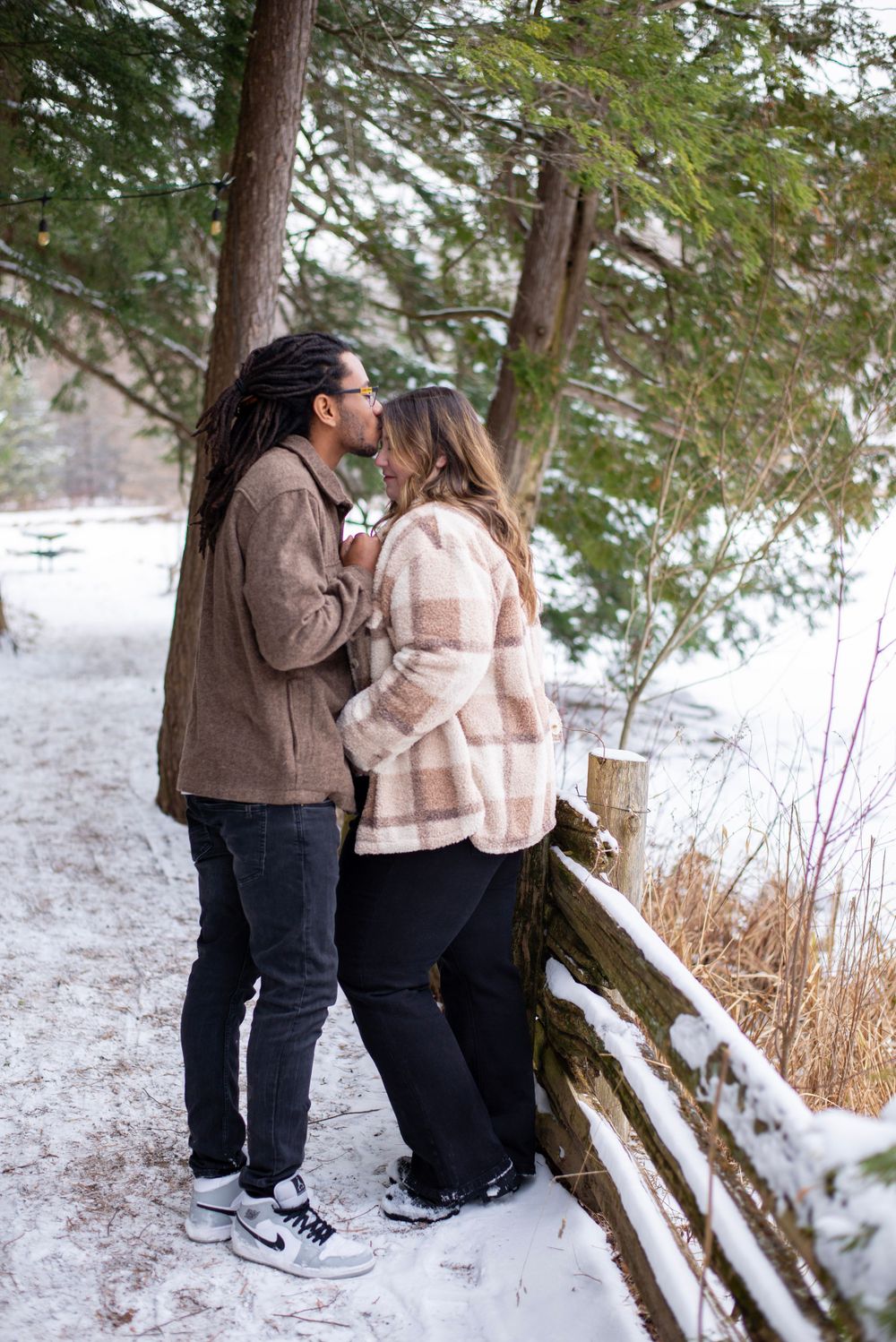 Couple sharing an intimate moment on a snowy winter trail beside evergreen trees and a wooden fence.