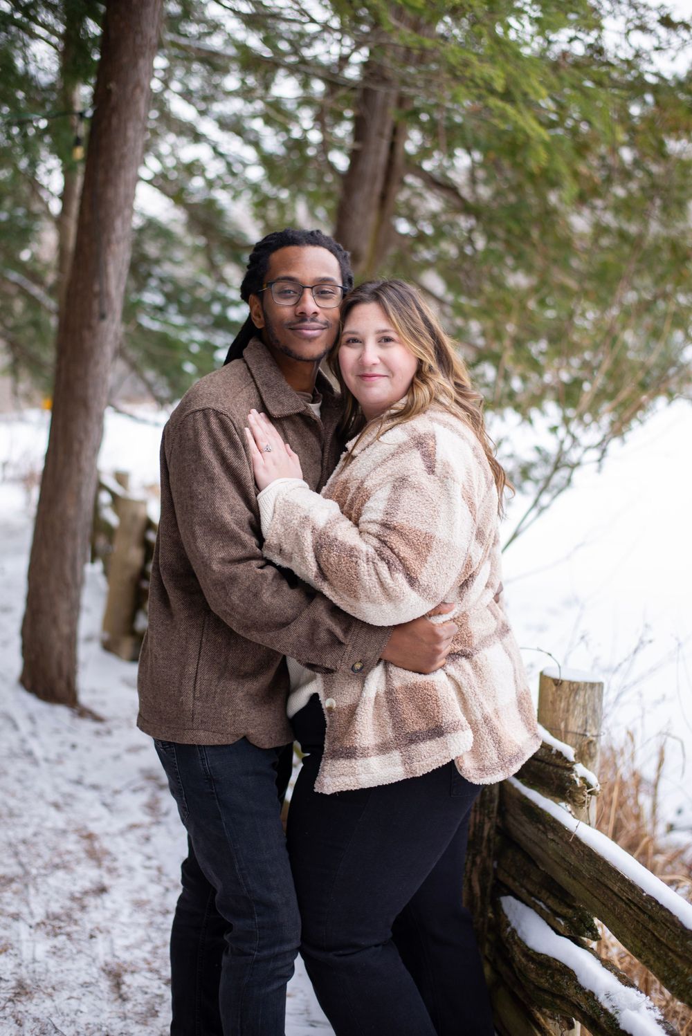 A couple embraces while wearing cozy sweaters in a snowy forest setting.