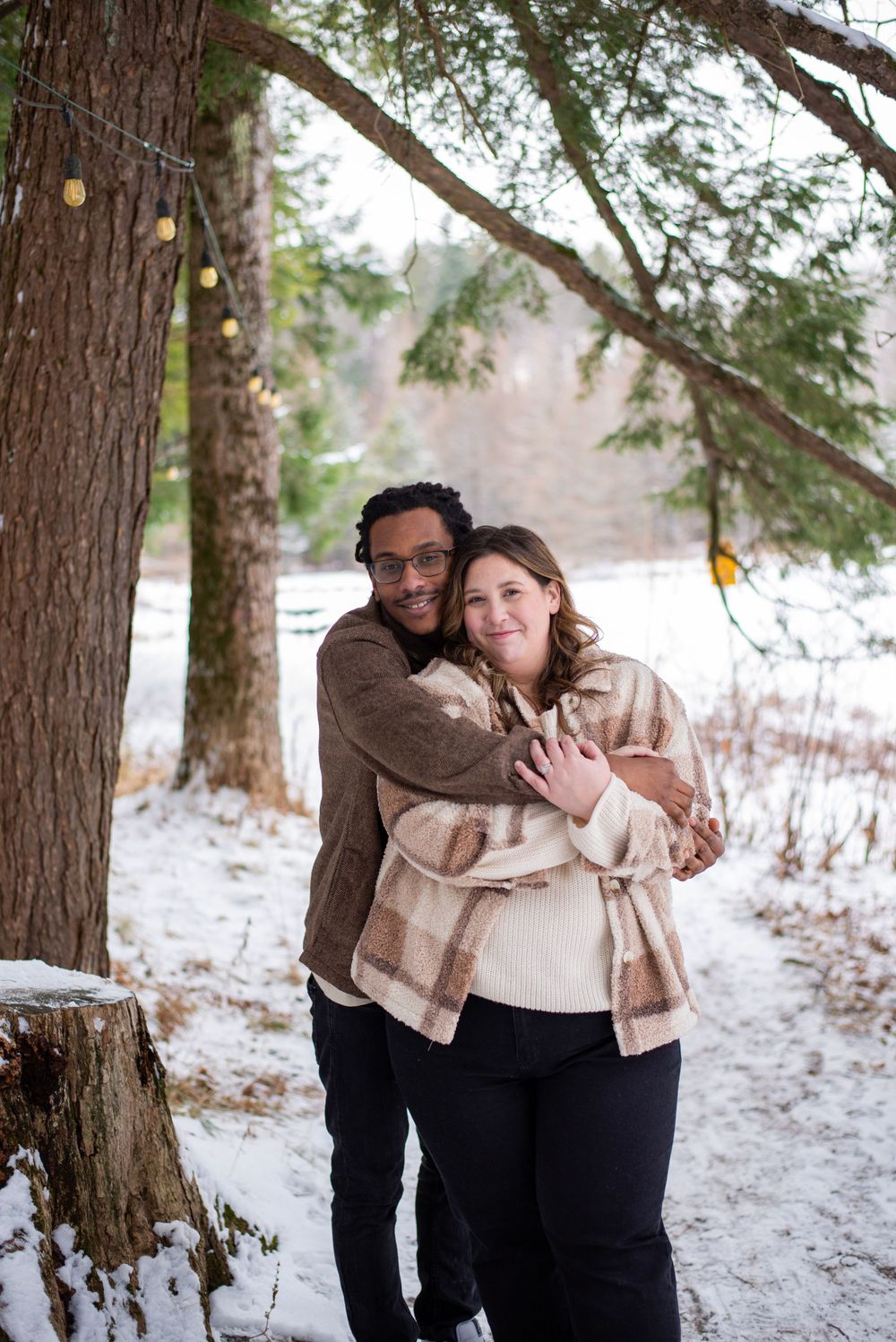 A couple embraces in a snowy winter forest setting while wearing cozy neutral-toned sweaters and jackets.