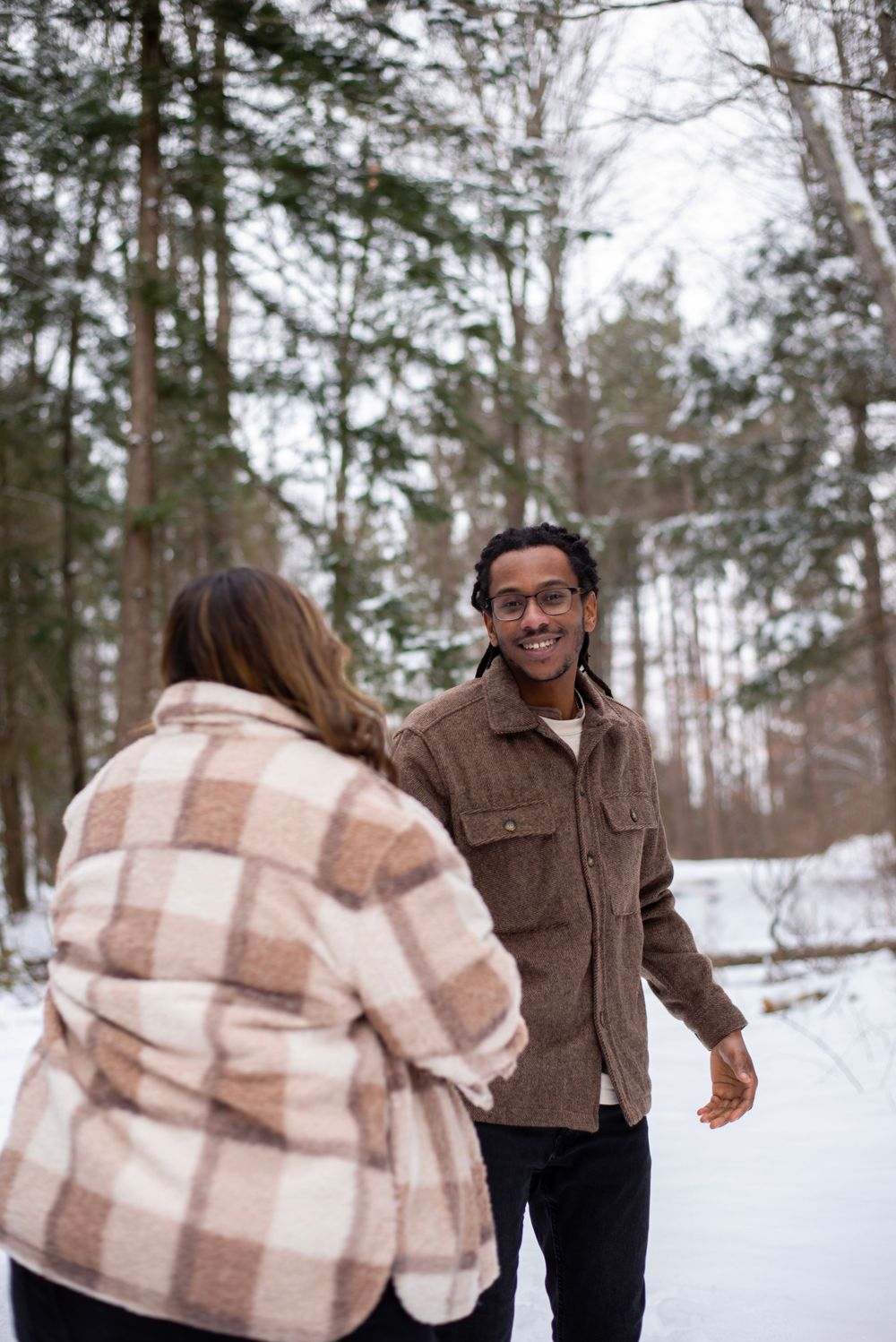 Two people in winter coats enjoying a snowy day in the woods, with evergreen trees in the background.