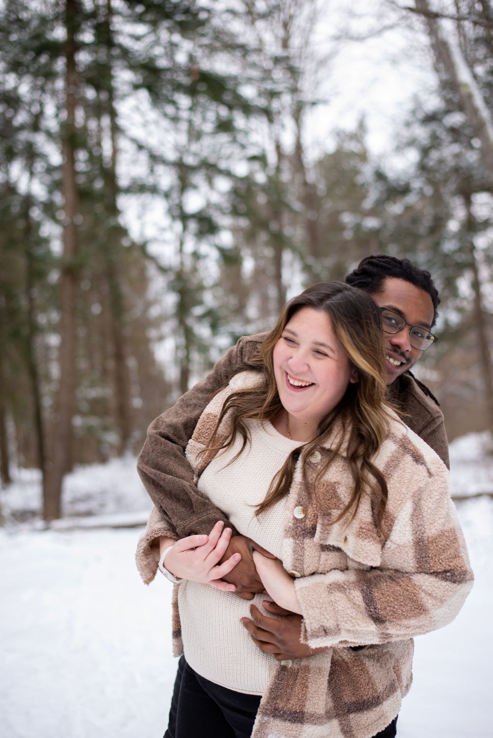 A couple embraces and laughs together on a snowy winter path surrounded by tall pine trees in the forest.