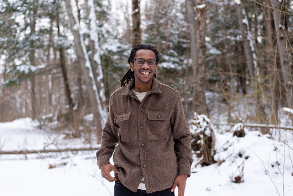 Person in brown fleece jacket stands smiling in snowy winter forest setting.