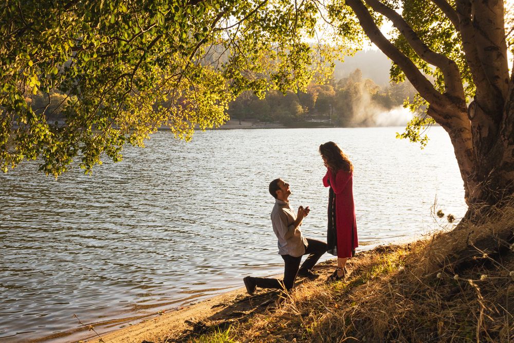 Couple sits together by a tranquil lake during golden hour with trees framing the scene.