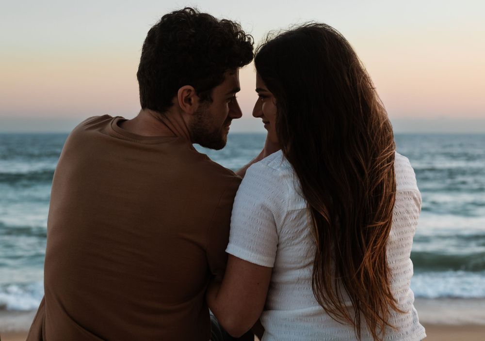 Silhouettes of a couple against a sunset ocean backdrop in matching white and brown outfits.