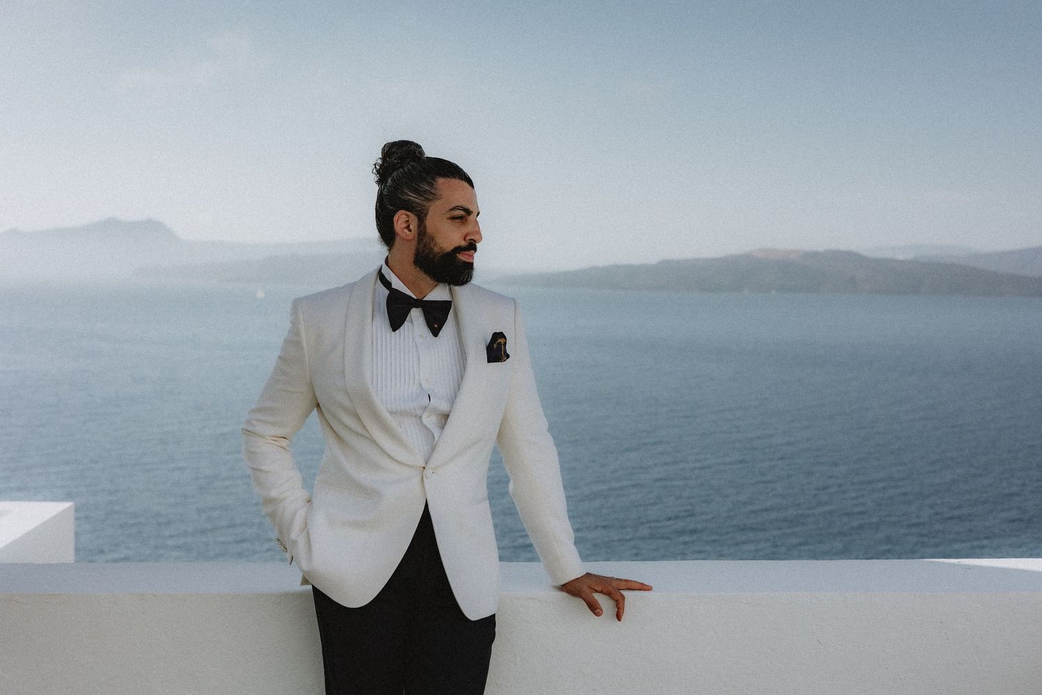 Groom in white tux standing against a white wall with the ocean behind in Santorini