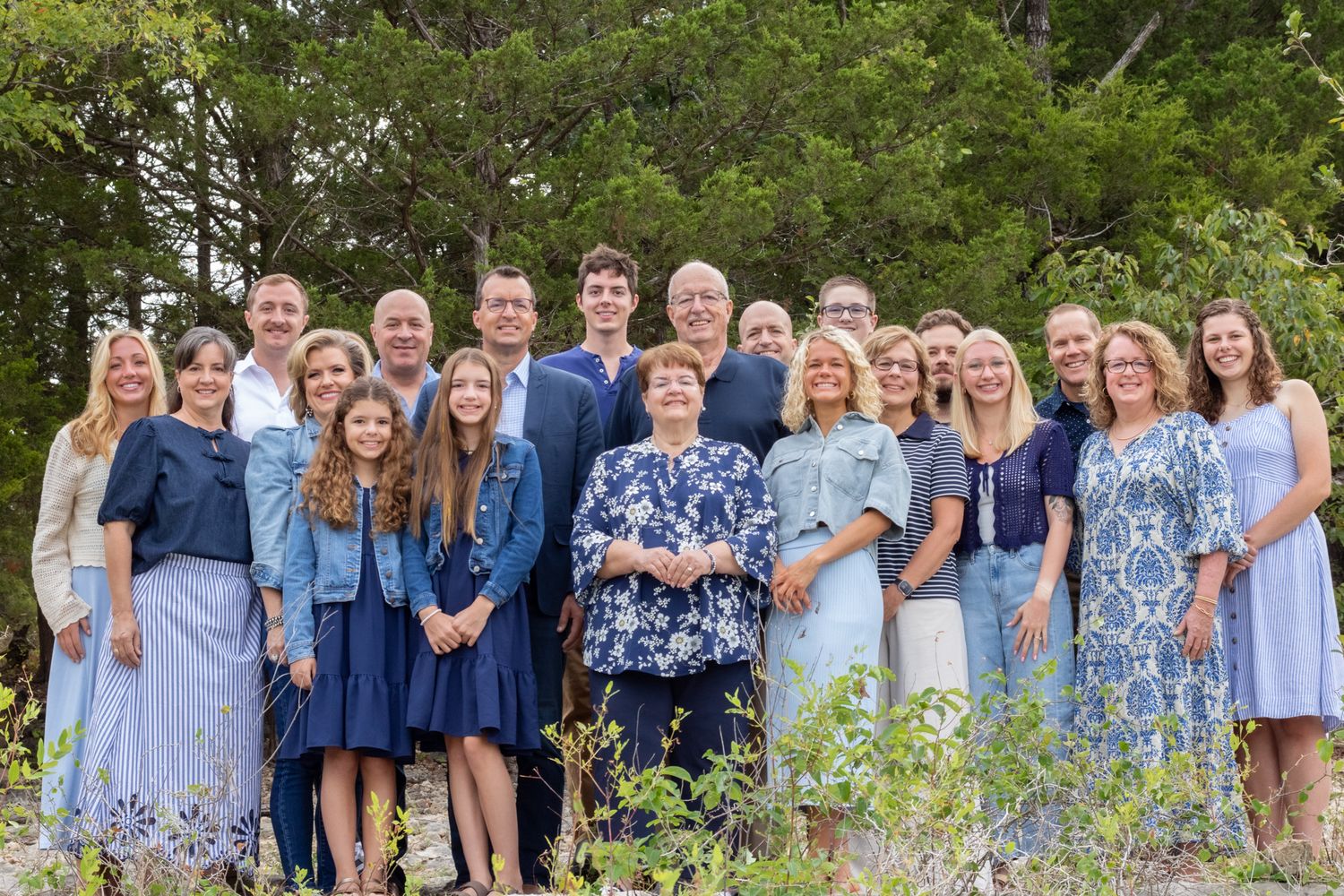 Family of 30 smiling looking at the camera in front of trees.