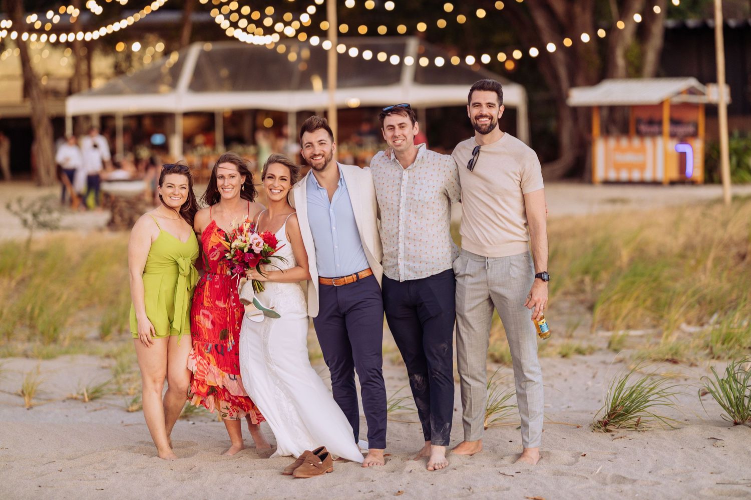 Group portrait on a beach at sunset with string lights glowing in the background.