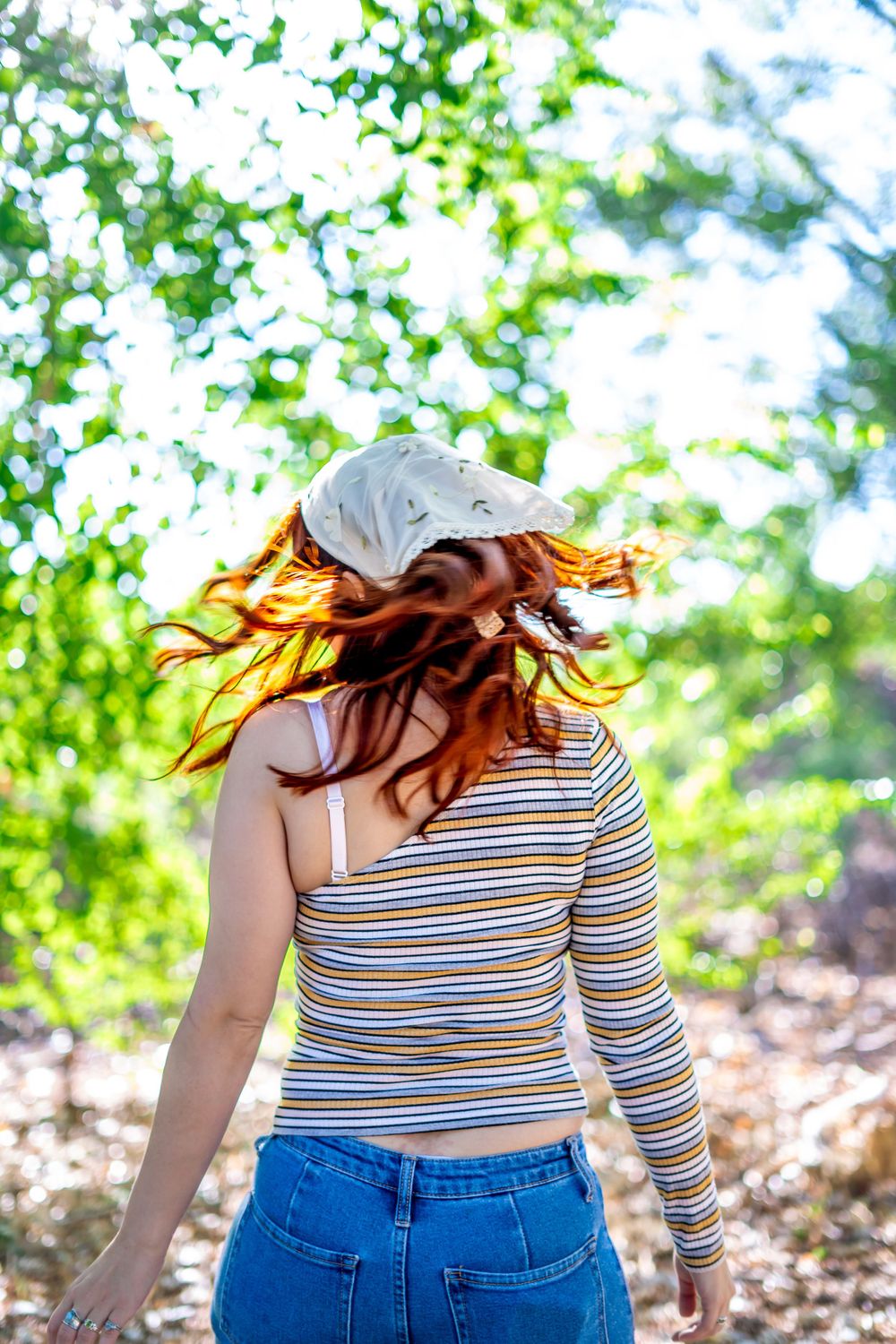 Back view of someone with long hair wearing a striped shirt and cap in a wooded area.