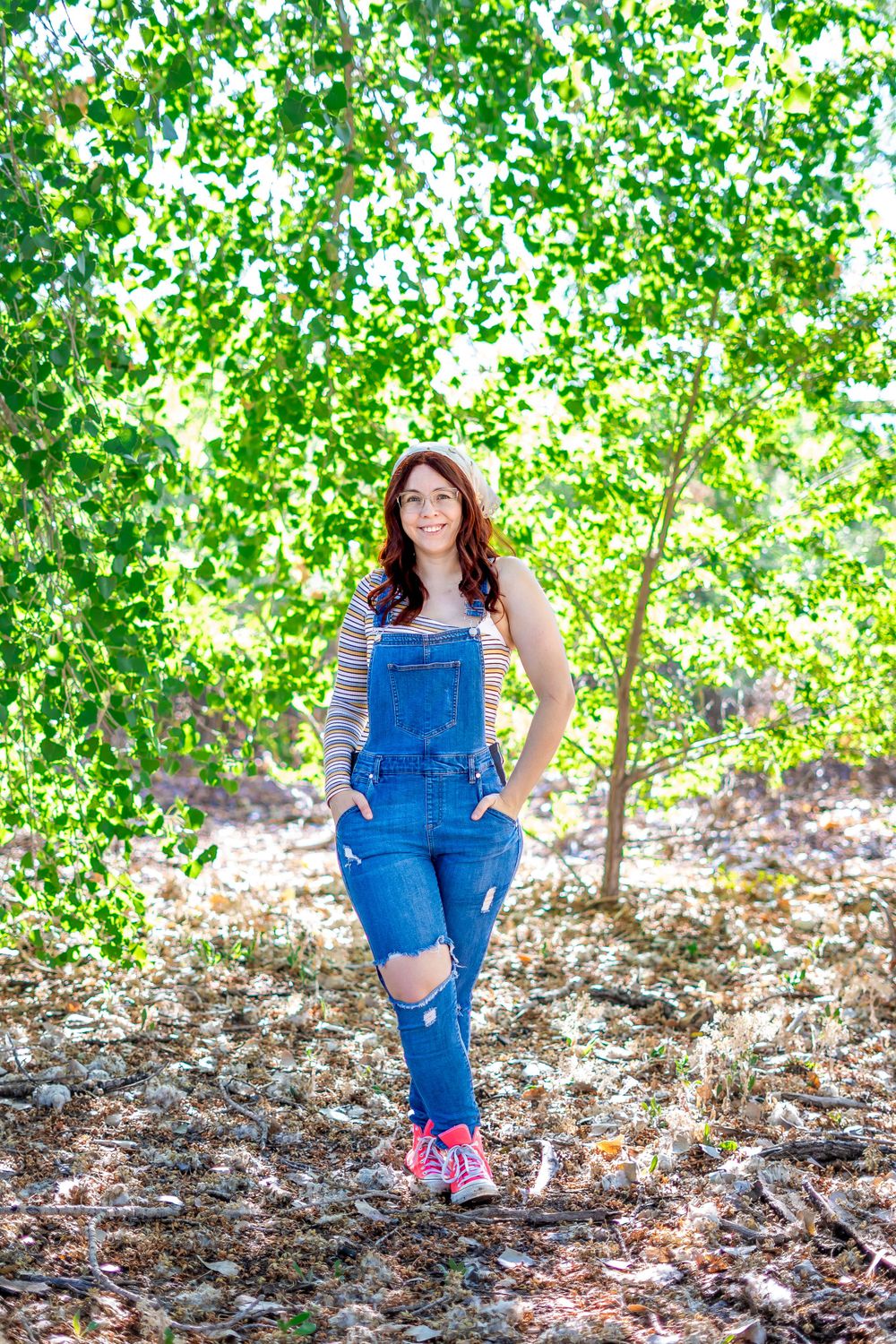 Full-length portrait in distressed denim overalls and striped top against trees.