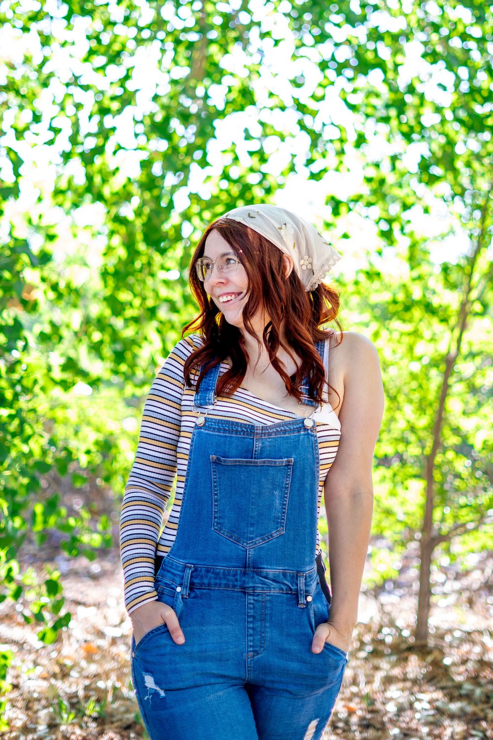 Casual outdoor portrait in denim overalls and striped shirt against green forest backdrop.