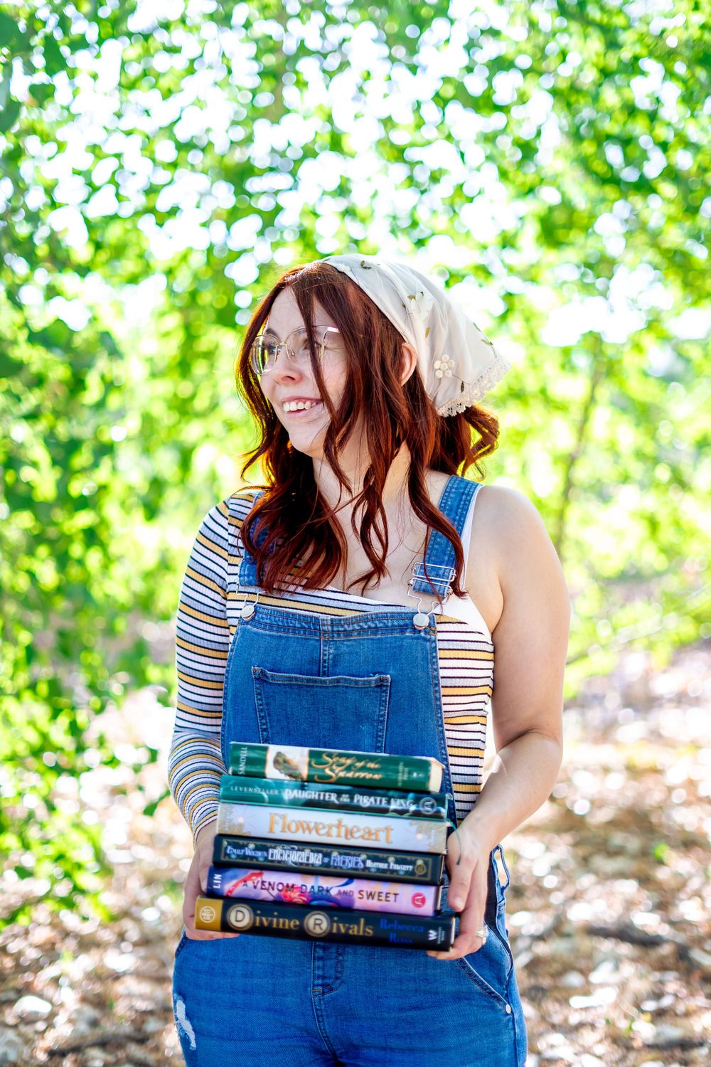 A candid shot in a forest setting showing someone in overalls carrying a stack of books against a leafy background.