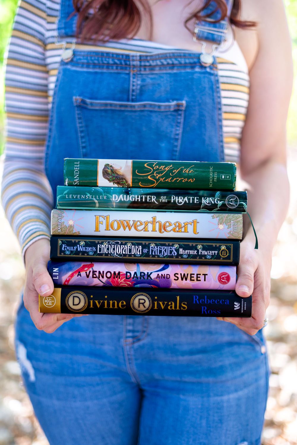 Stack of popular books held against blue denim overalls showing titles like Flowerheart and Divine Rivals.