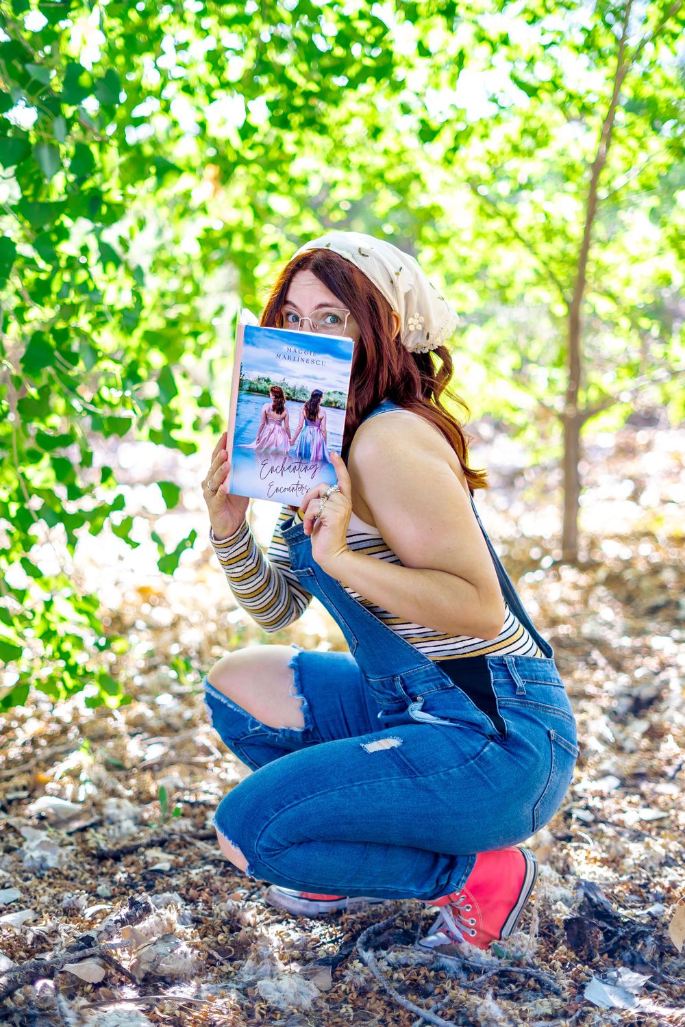 Playful pose holding a book while wearing overalls and red sneakers in a wooded setting.