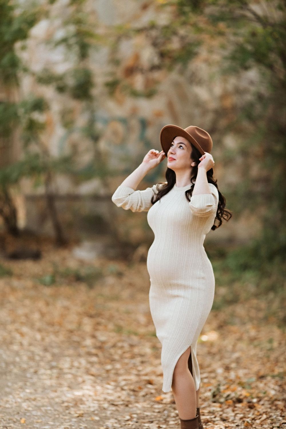 A woman in a cream sweater dress and brown hat poses on an autumn path.
