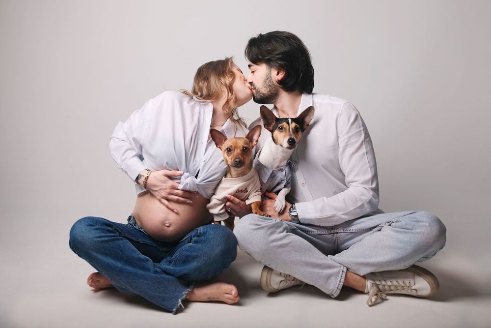 Studio maternity photo with couple in casual attire sitting with their dog.