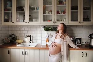 Maternity photo of woman relaxing in modern white kitchen with glass cabinets.