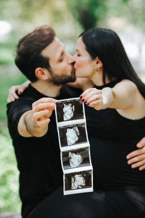 Couple shares intimate moment while holding ultrasound photos outdoors.
