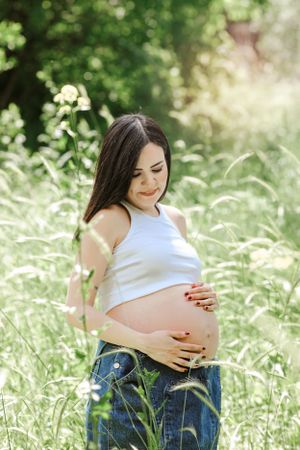 Expectant mother in white top touches her belly in sunny meadow.