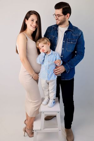 Family portrait with child standing on white step stool against neutral background.