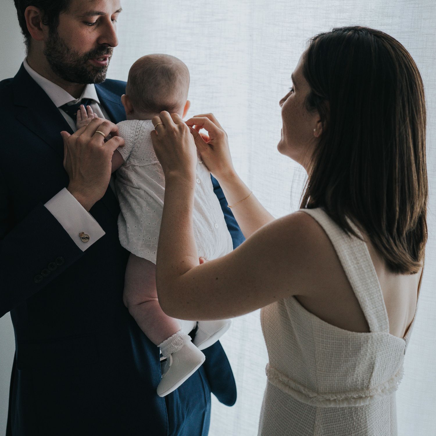 Couple in formal attire tenderly caring for their baby, man in navy suit, woman in white dress, soft natural light.