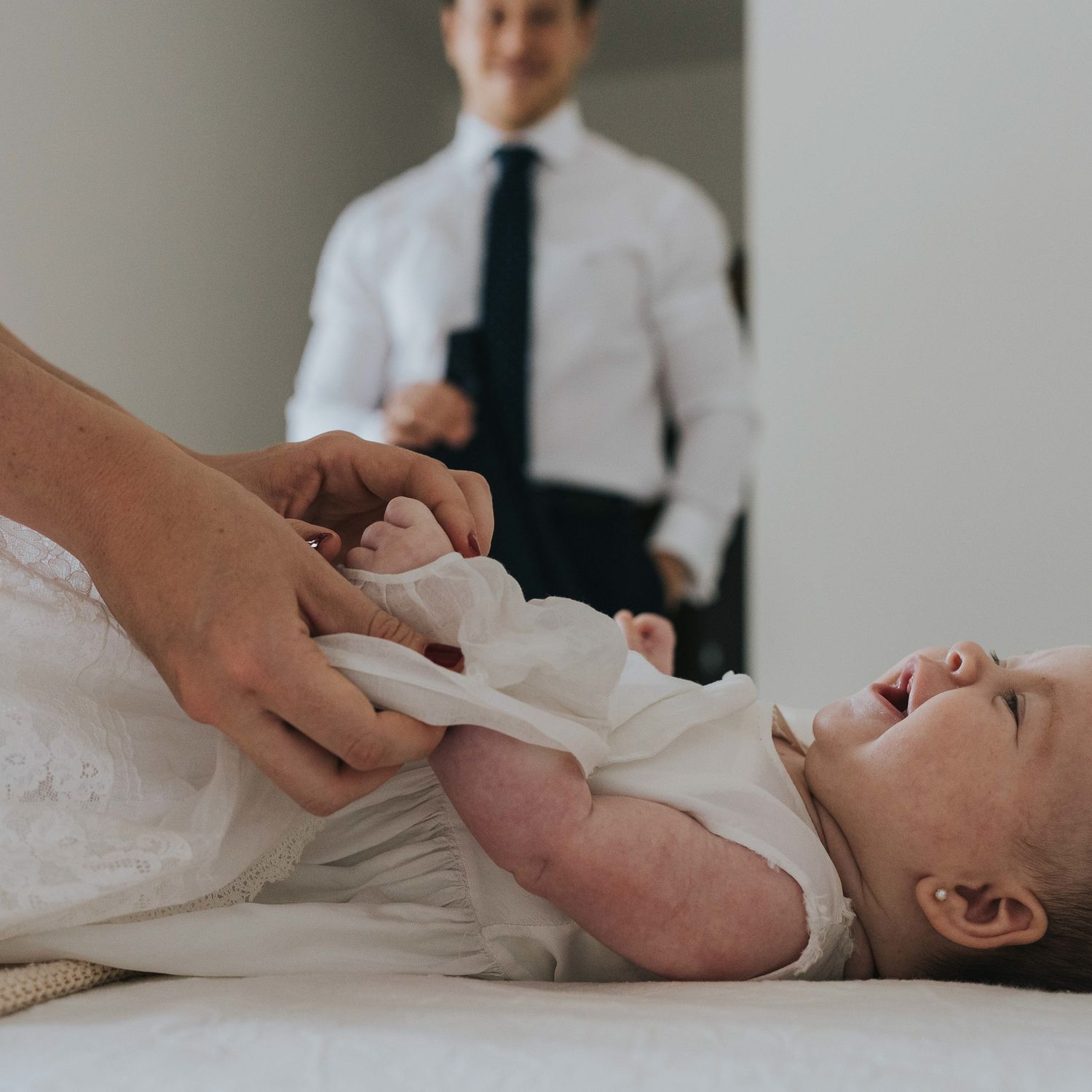 A baby in a white dress lies on a changing table while an adult holds their hands, with a man in a suit visible behind.