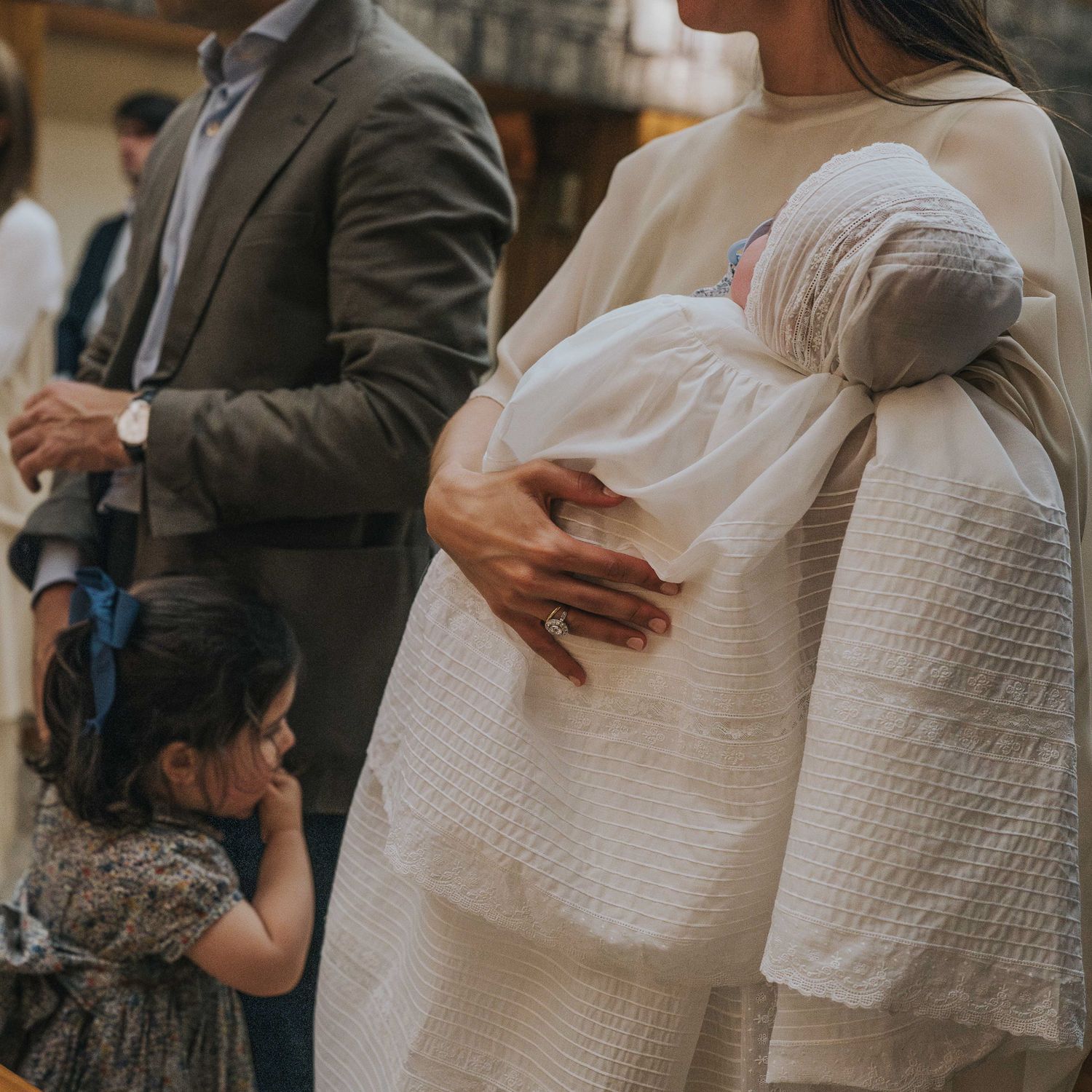 A mother holds a newborn in white christening gown at baptism ceremony with toddler daughter and father nearby.