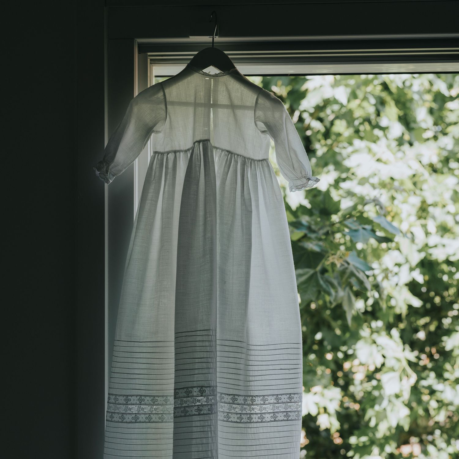 A delicate white christening gown hanging on a hanger in front of a sunlit window with lush green trees.