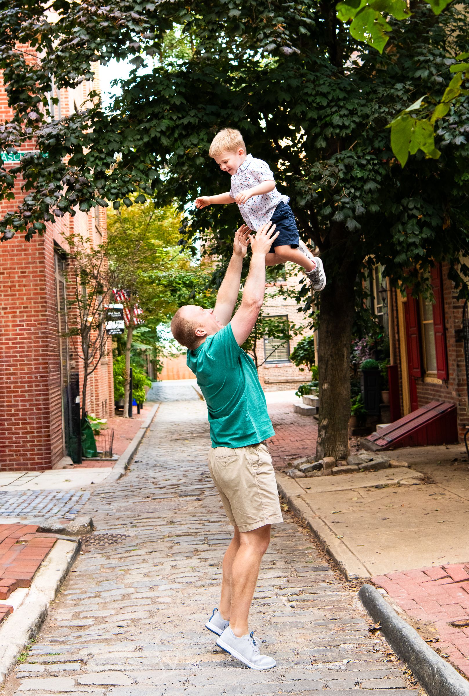 Dad in a green shirt lifting and playing with his son on the historic Philadelphia cobblestone street.