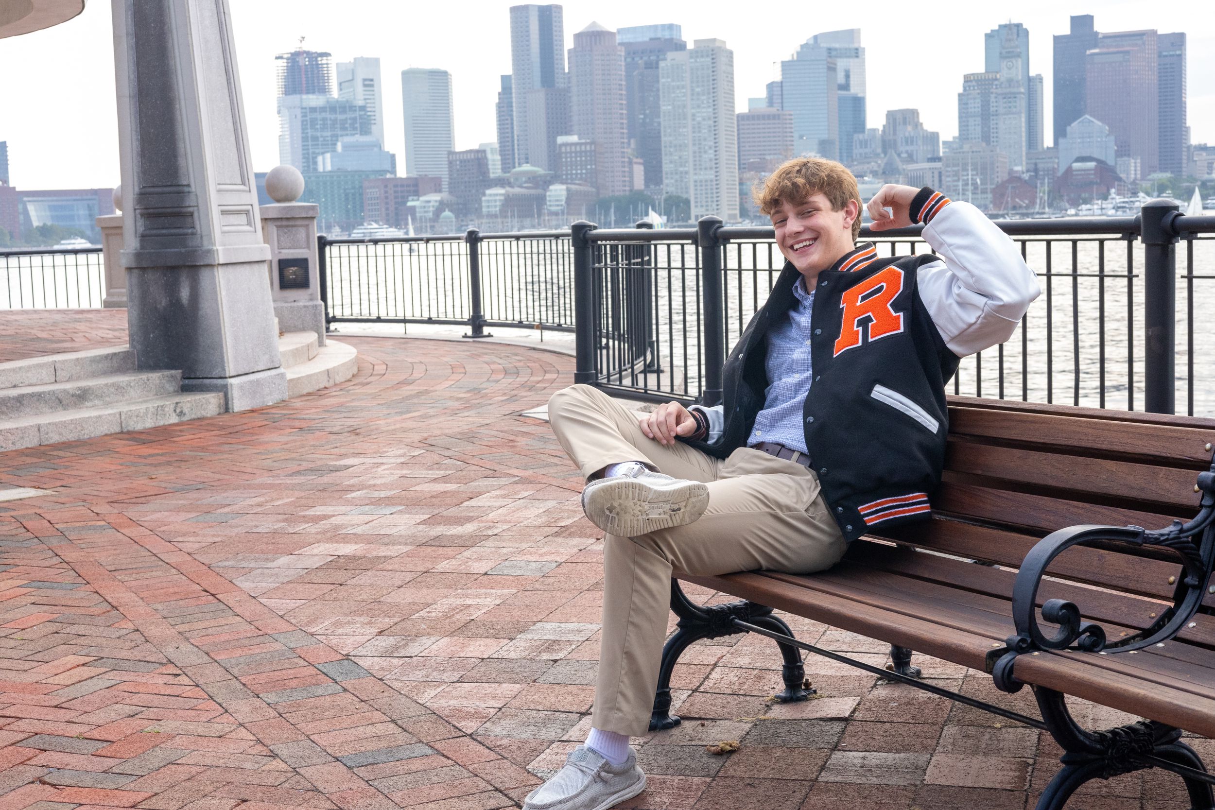 Someone in varsity jacket reading on bench with Boston harbor skyline in background.