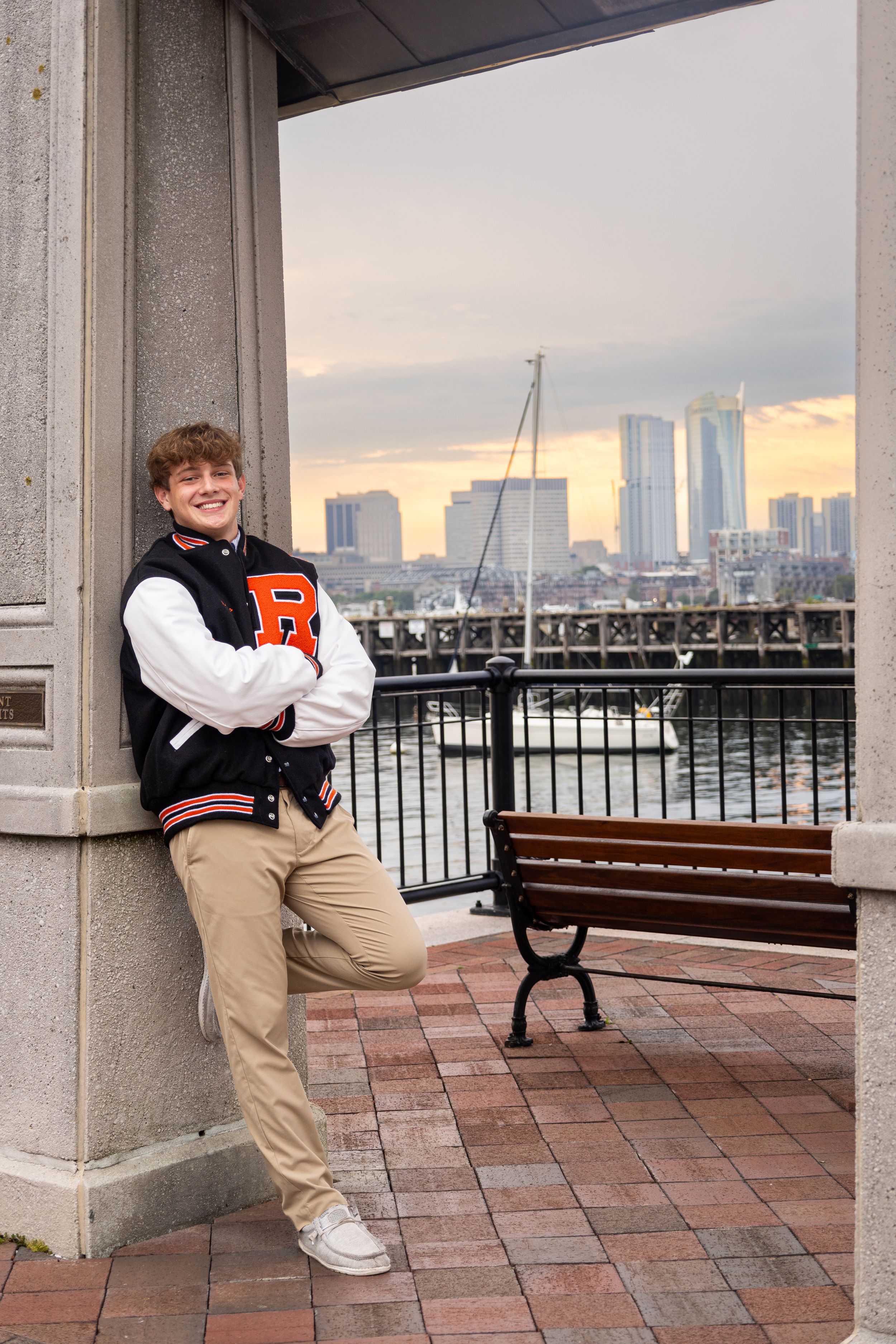 Student in varsity jacket poses against column with Boston harbor and sunset in background.