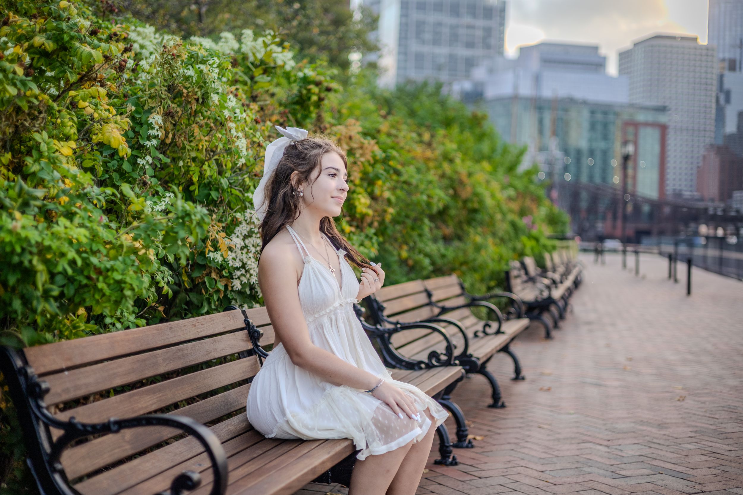 A person in a white dress sits on a park bench with city buildings in the background.