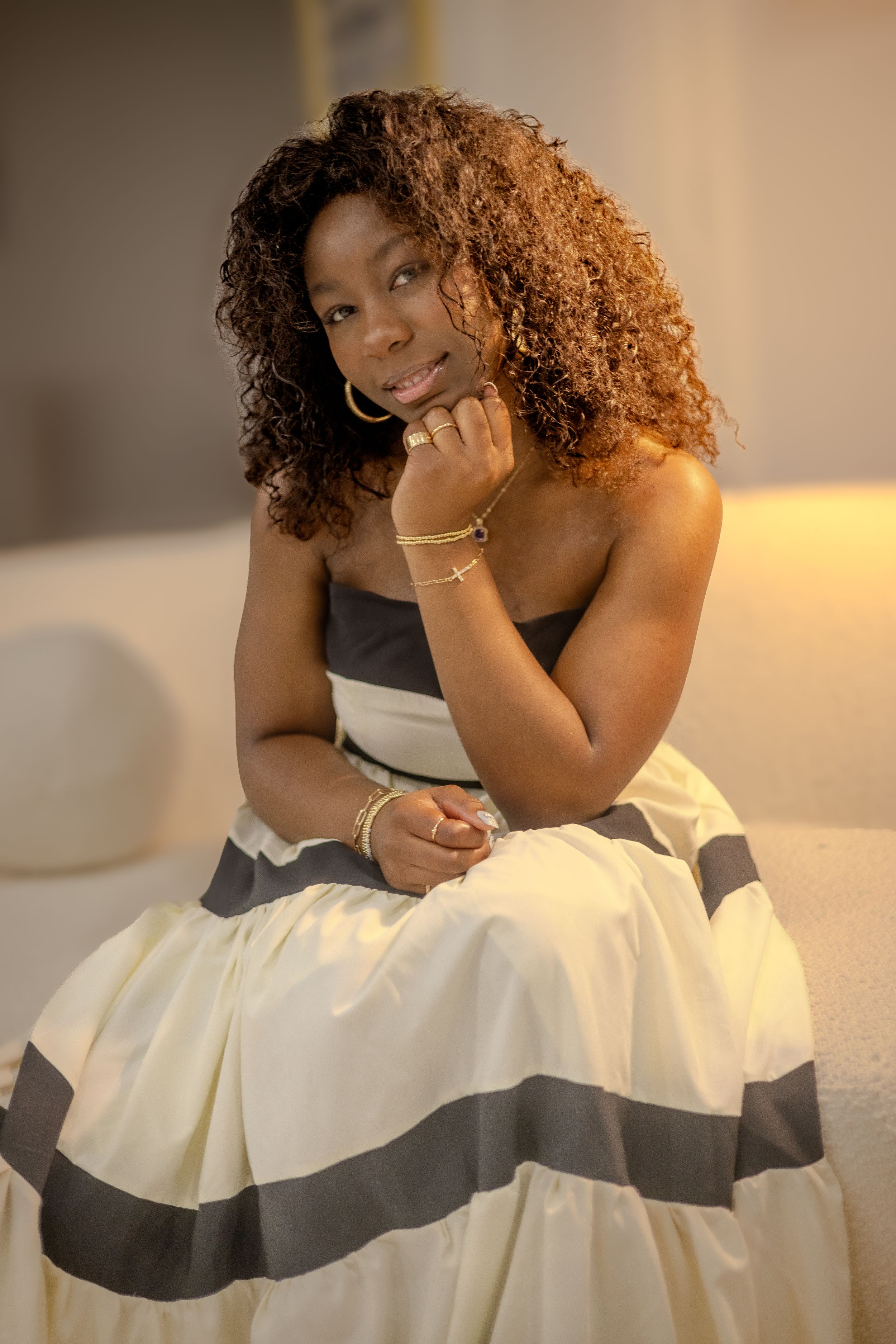 A model poses in an elegant strapless white gown with black stripes while sitting gracefully on a couch.