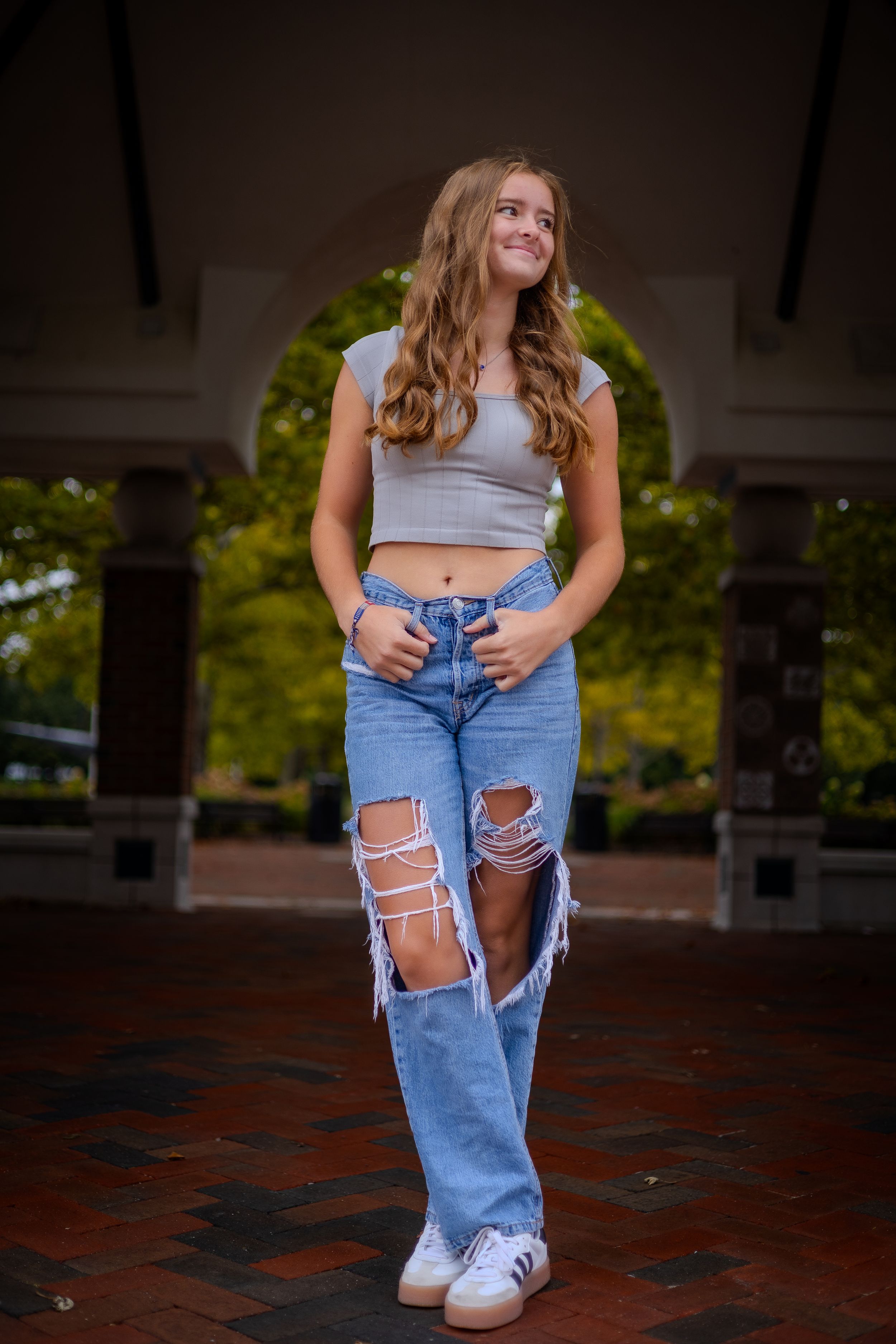 Portrait shot of a woman in distressed jeans and gray crop top posing in arched brick courtyard.