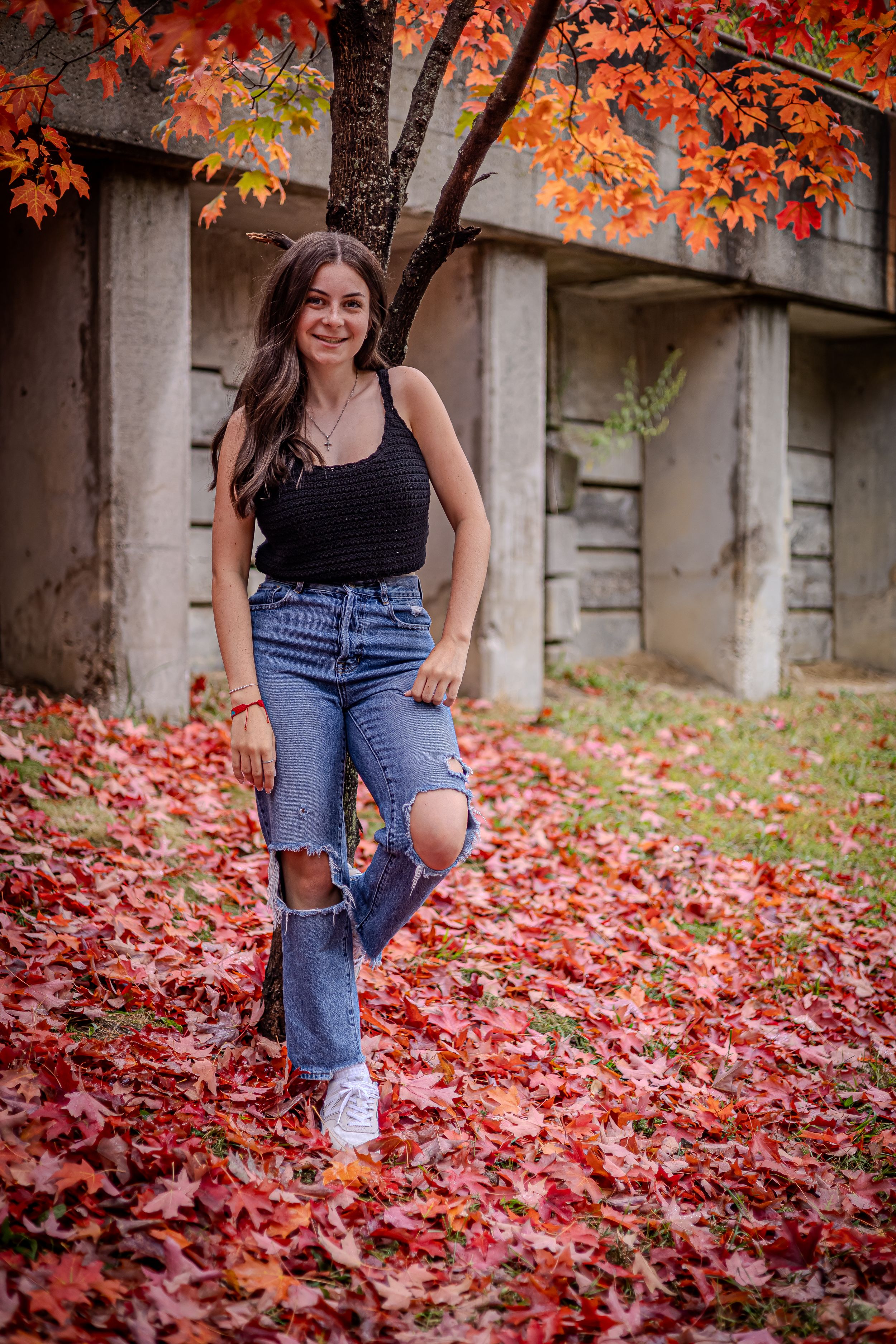 A person in ripped jeans and black top poses outdoors among vibrant red fall foliage and autumn leaves.