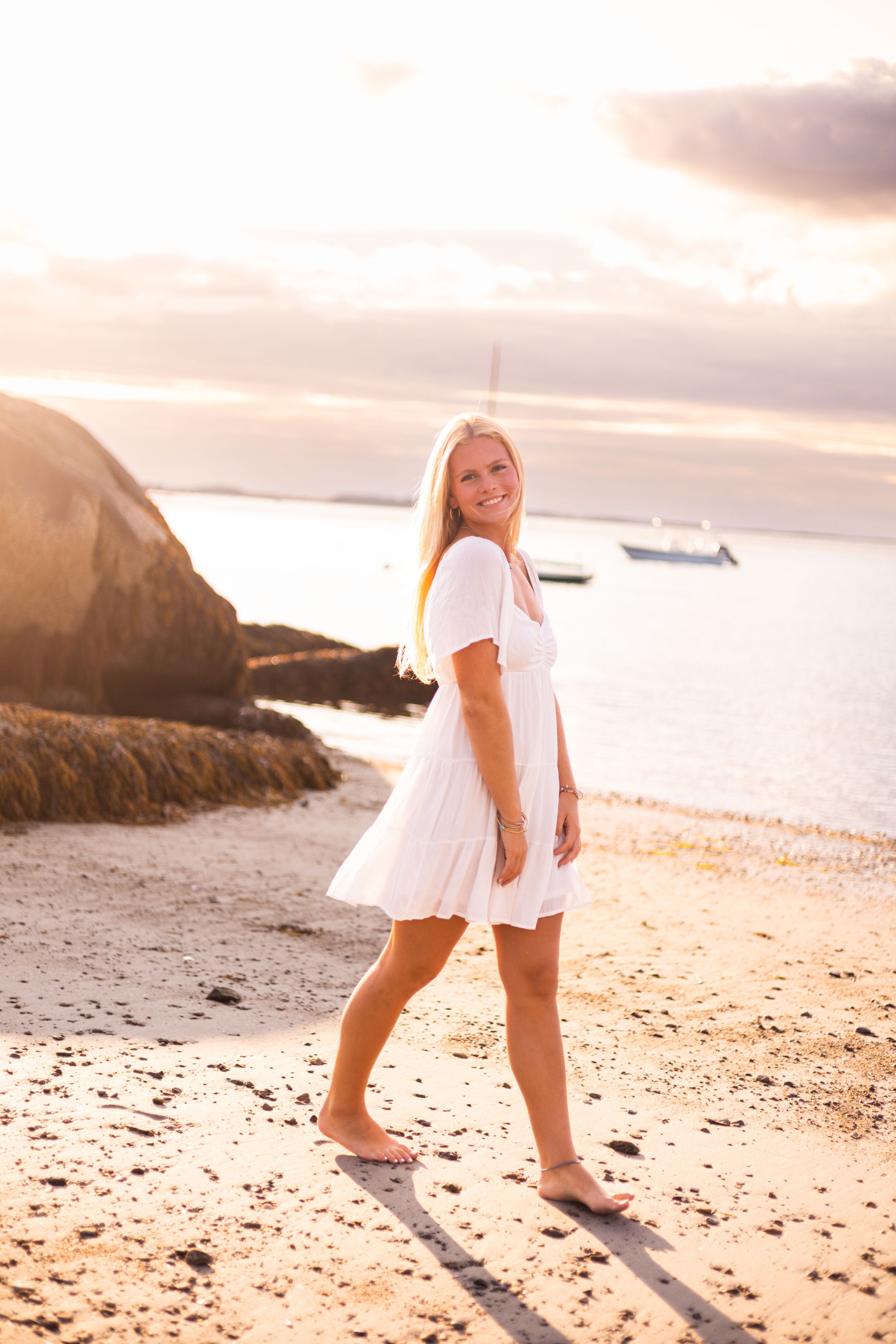 Someone in a white dress walking barefoot on beach at sunset with rocks and ocean in background.