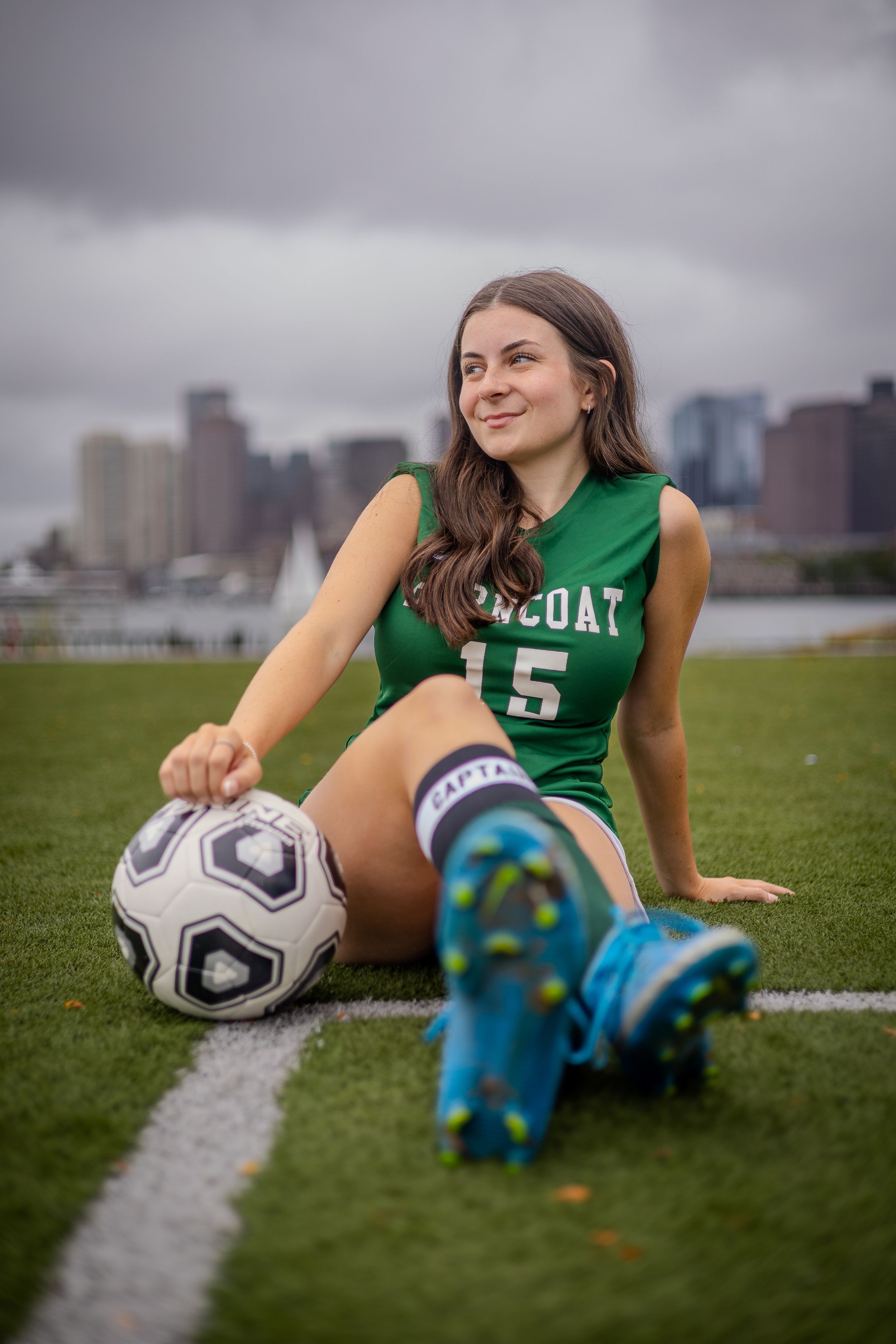 Soccer player in green jersey sits on field with ball and cleats against city skyline backdrop on cloudy day.