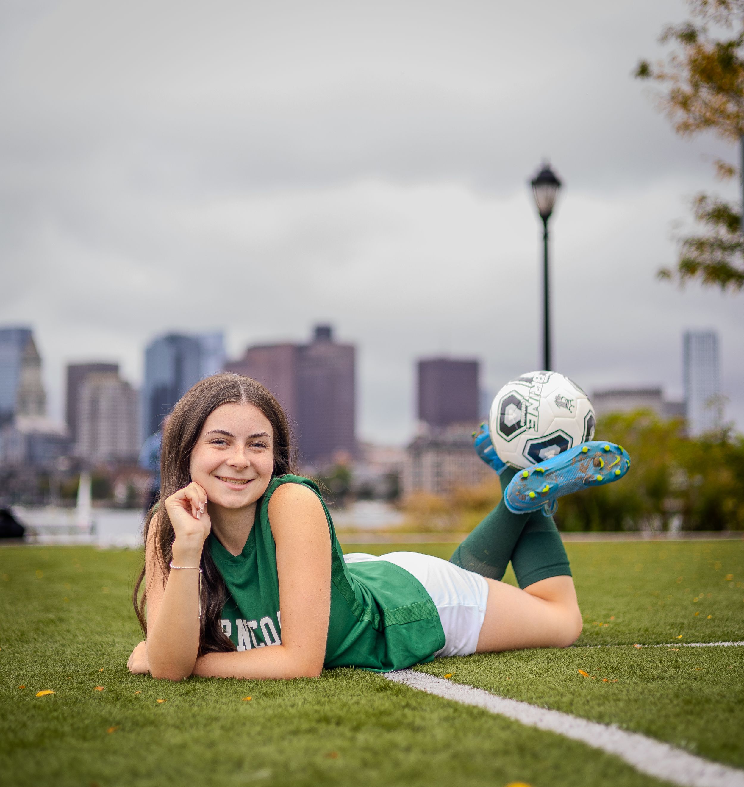 Soccer uniform clad player lies on grass field with skyline and lamp post in background.