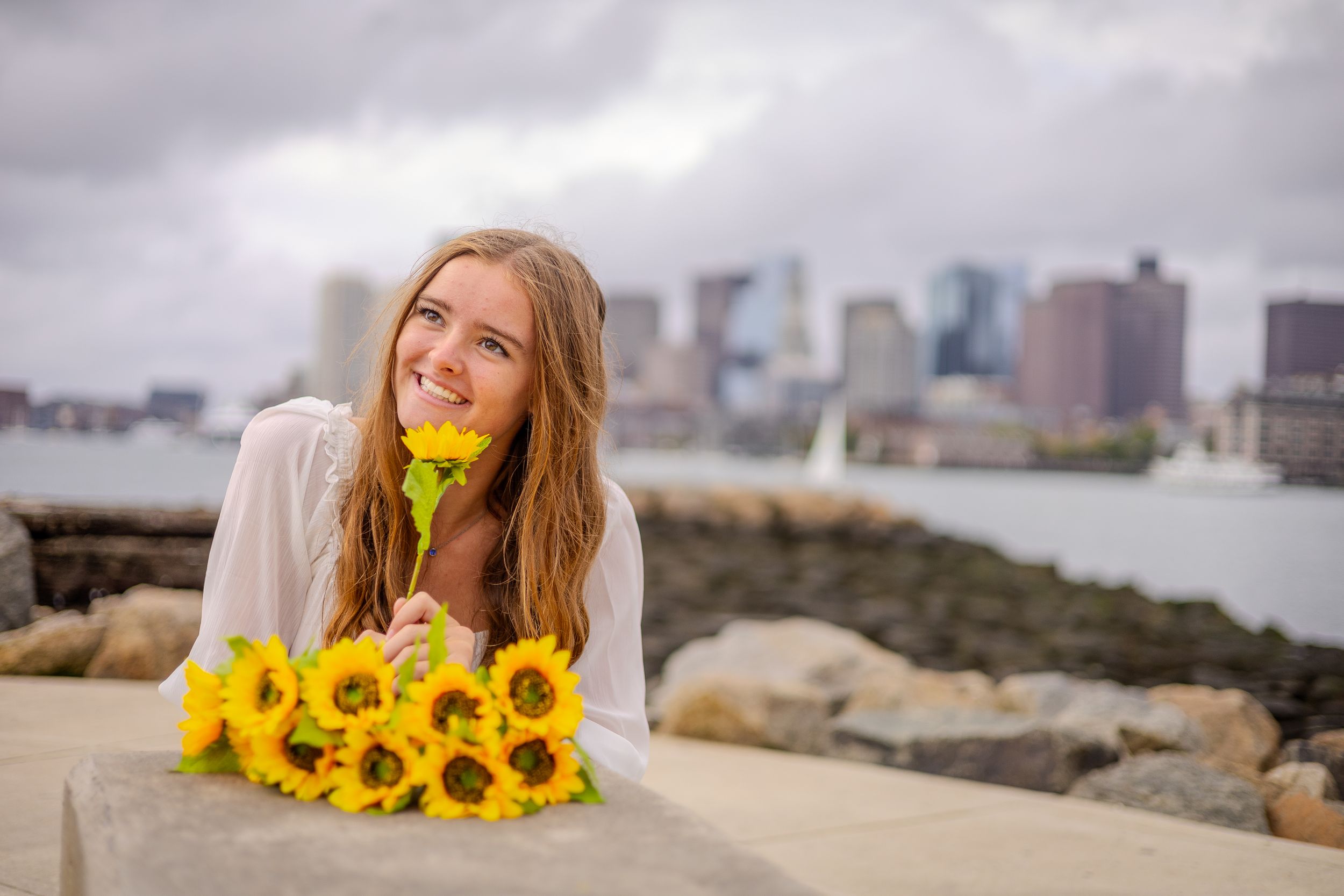 Young woman poses with bright yellow sunflowers along a rocky waterfront with a city skyline in the background.