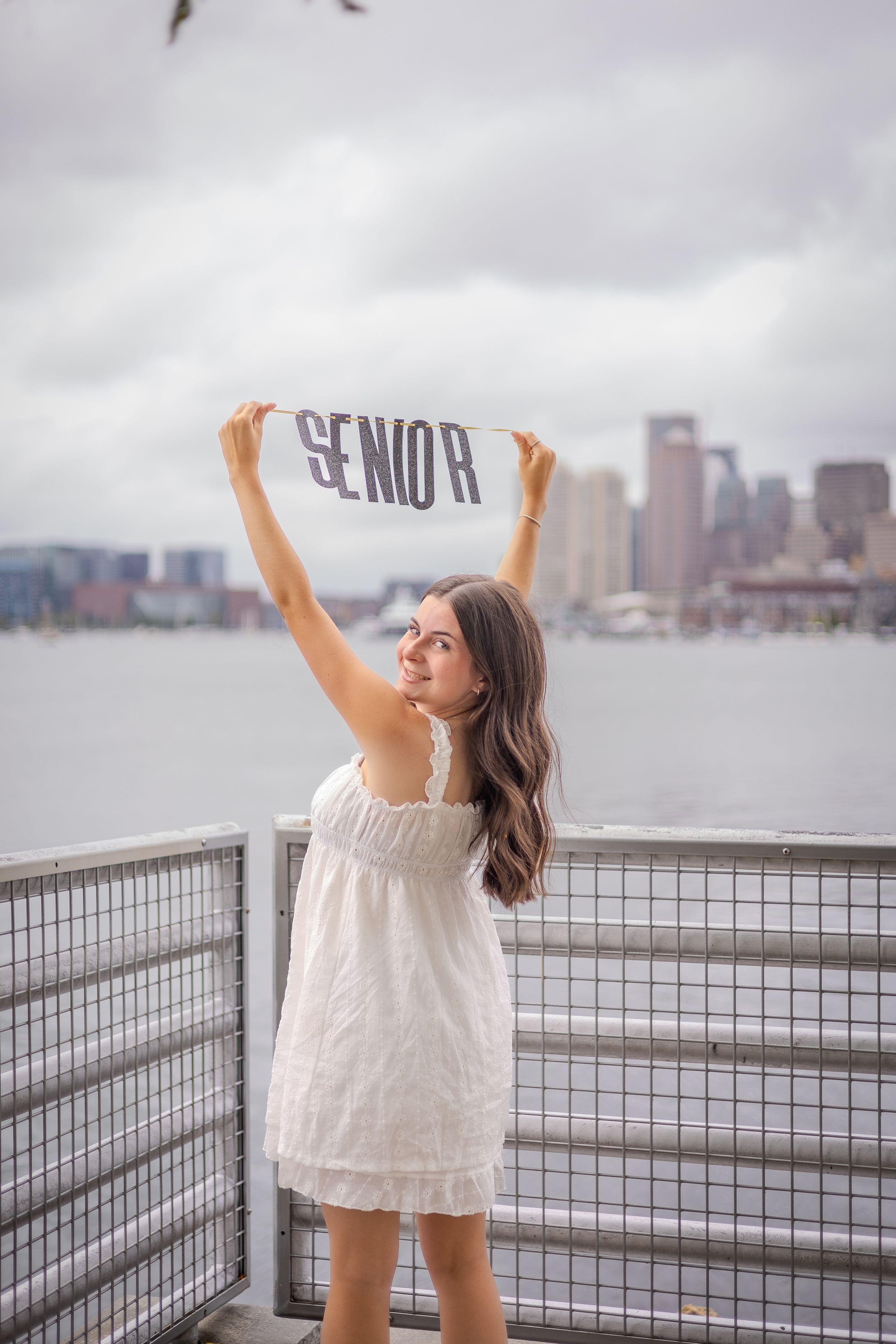 Figure in white dress raises 'SENIOR' sign against city skyline backdrop near waterfront railing on cloudy day.