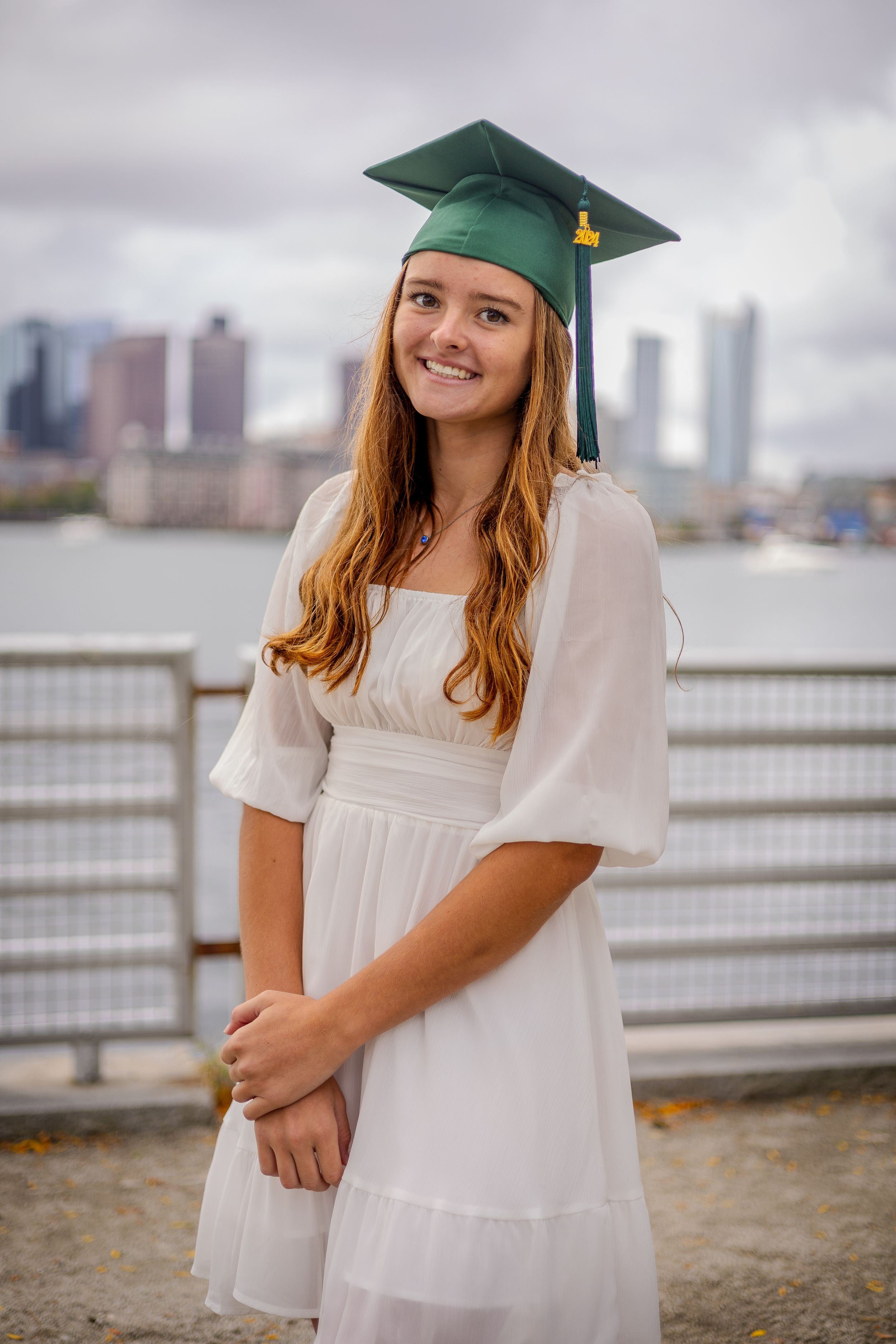 Graduate in white dress and green cap poses by waterfront with city skyline in background.