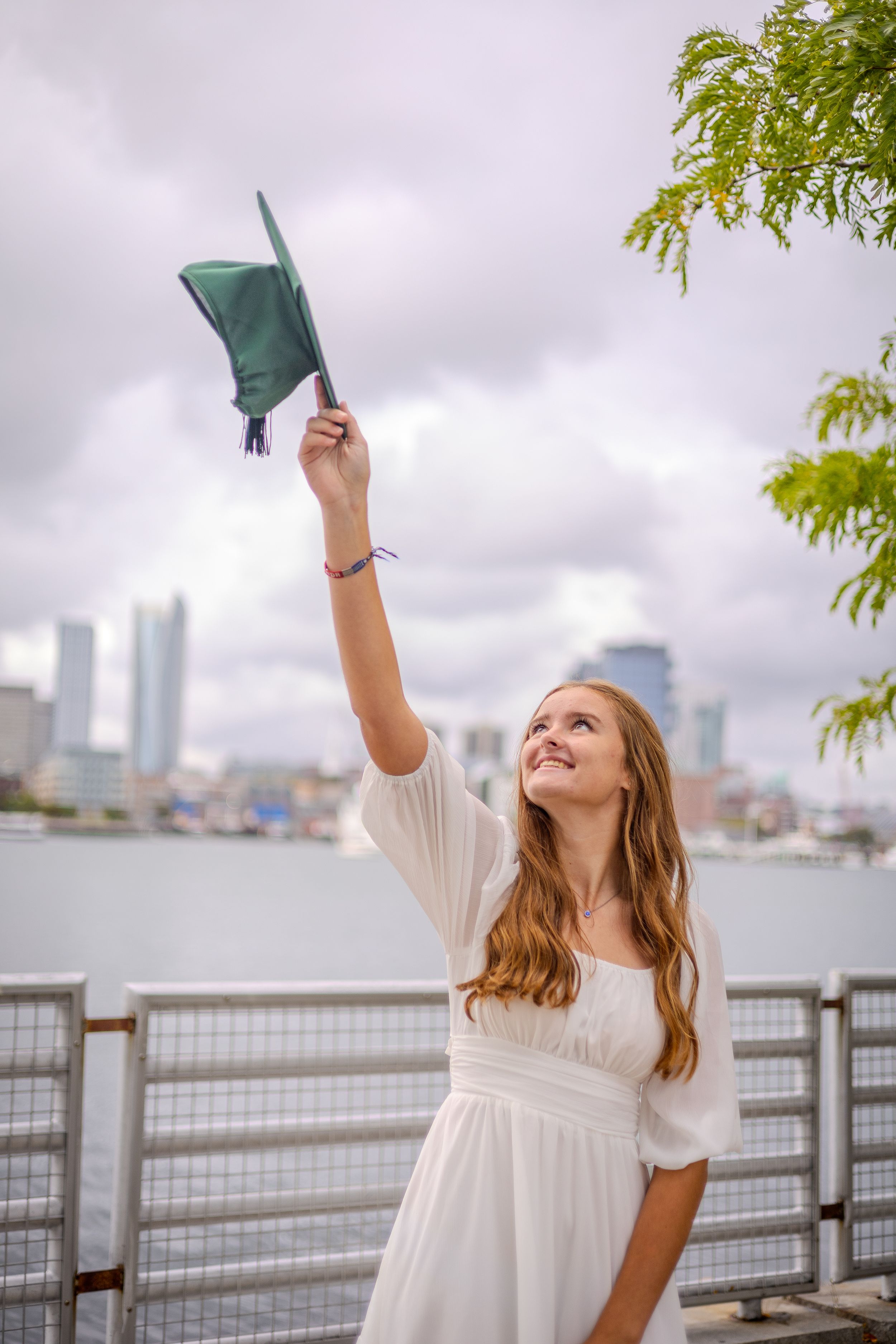 A graduate in a white dress raises their graduation cap by the waterfront with a city skyline in the background.