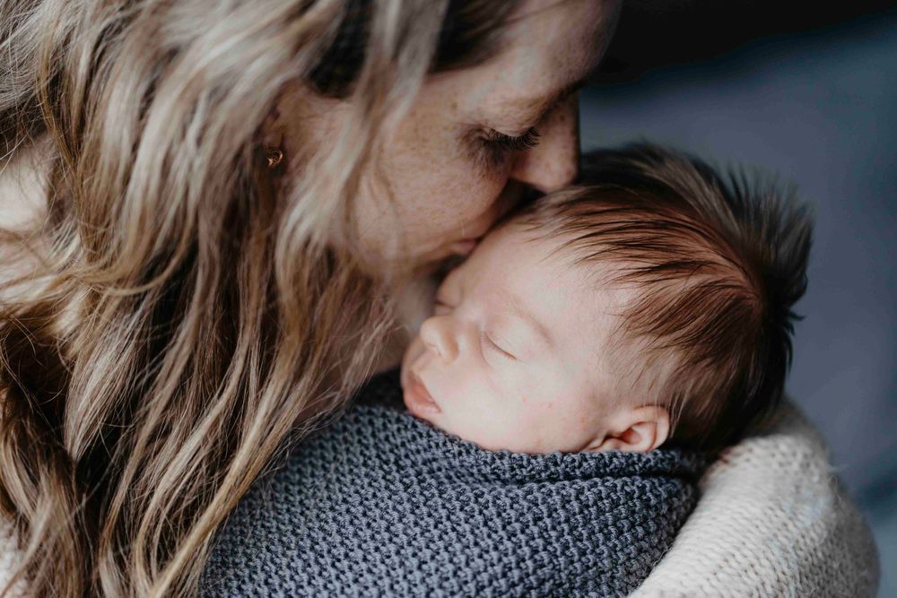 Mãe beija suavemente a testa do seu bebê adormecido, envolvido em manta azul, num retrato de amor e conexão profunda.