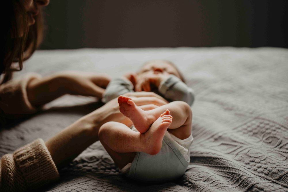 Bebê deitado na cama com a mãe ao lado, em um momento terno e íntimo de conexão no ambiente familiar.