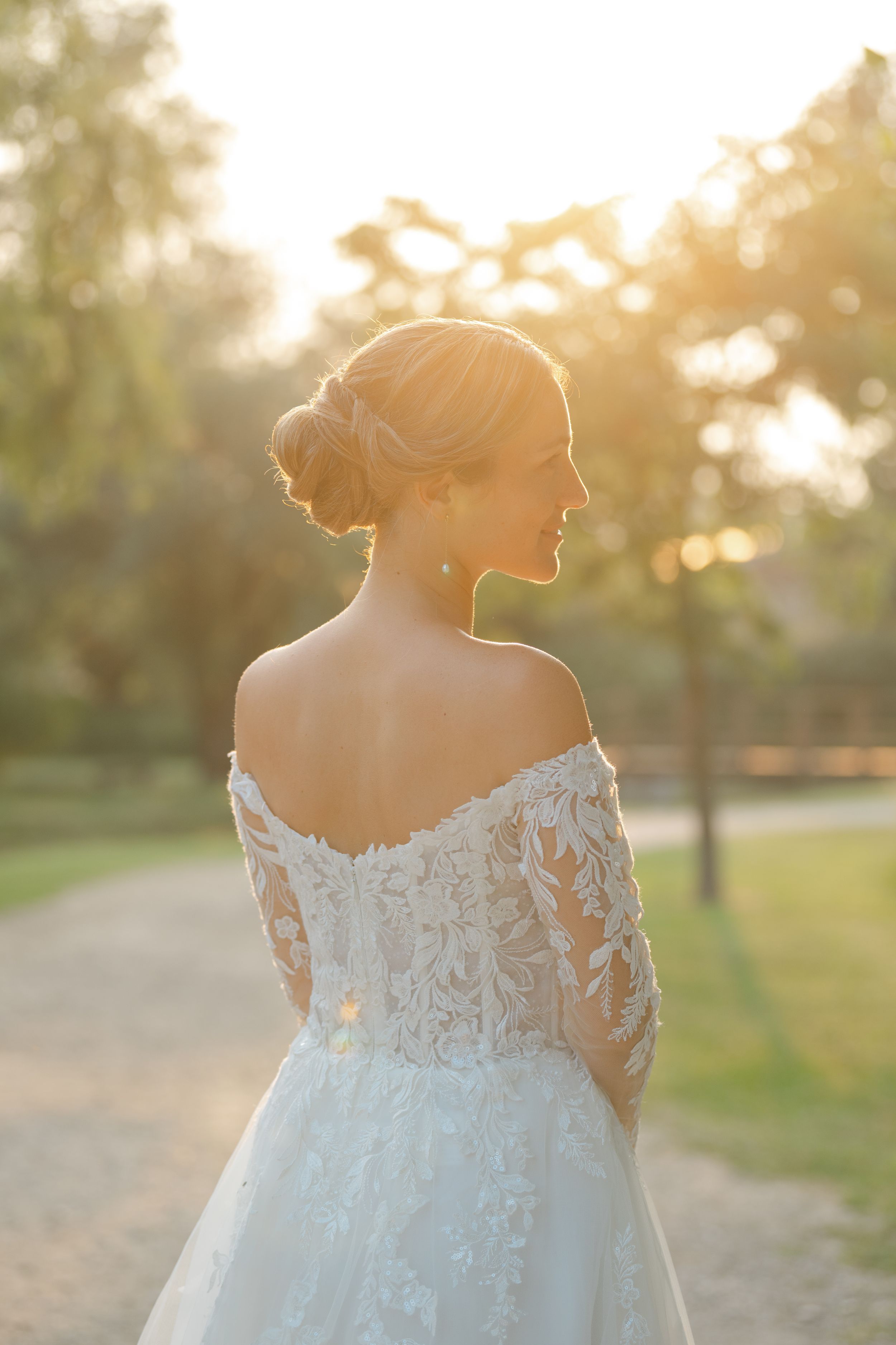 A bride in an off-shoulder lace wedding gown stands in golden sunlight against a blurred outdoor background.