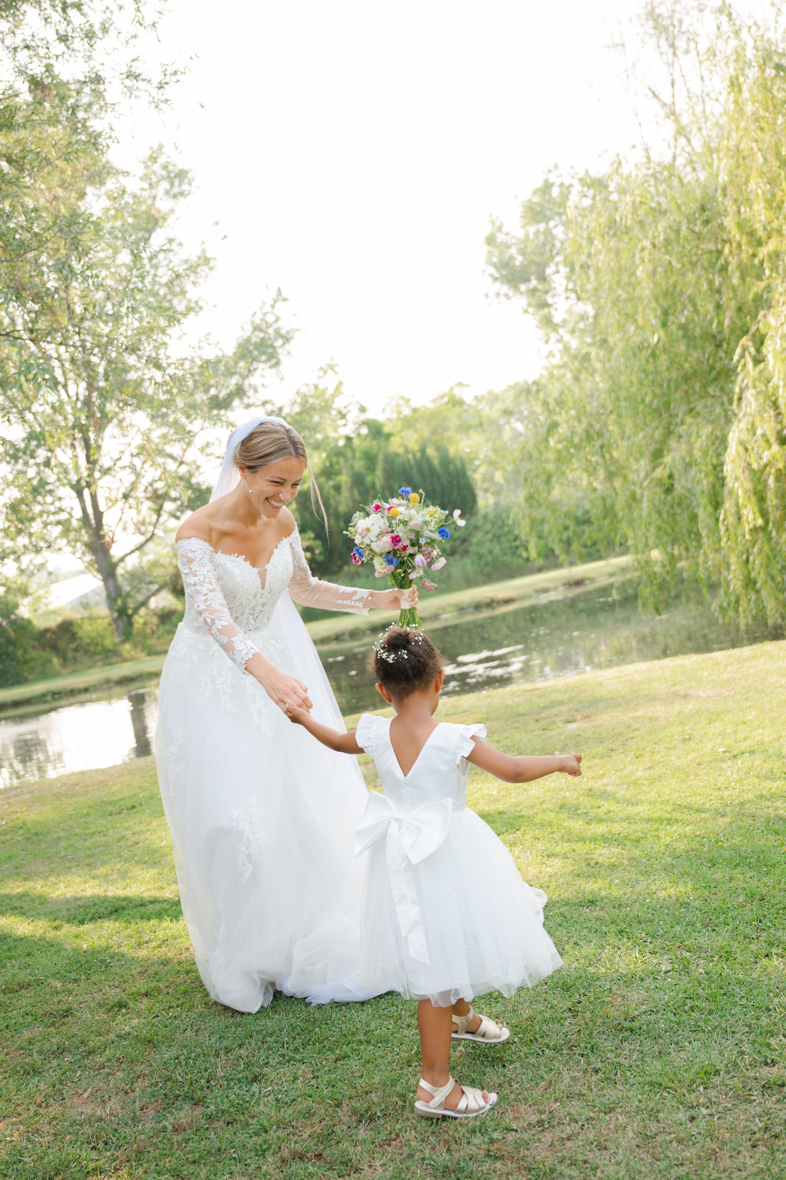 A bride sits on the grass in a white wedding dress and catches flower petals being tossed in the sunlit garden setting.