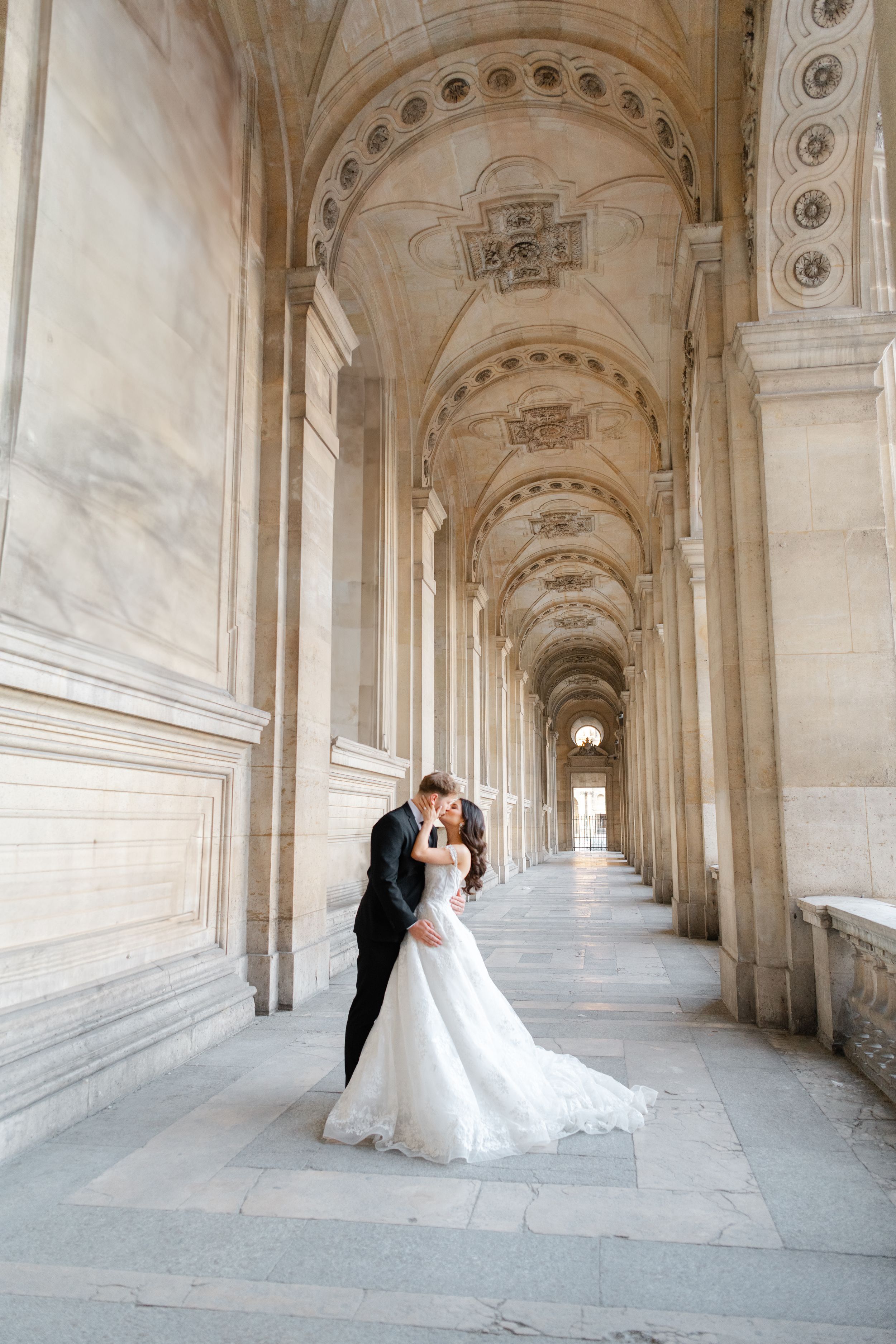 A couple in wedding attire embraces in an elegant arched hallway with classical architectural details.