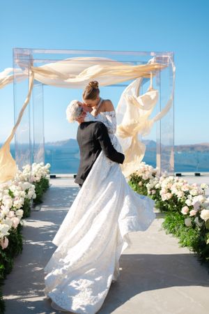 Wedding ceremony under a clear acrylic arch adorned with white flowers and flowing fabric by the ocean.