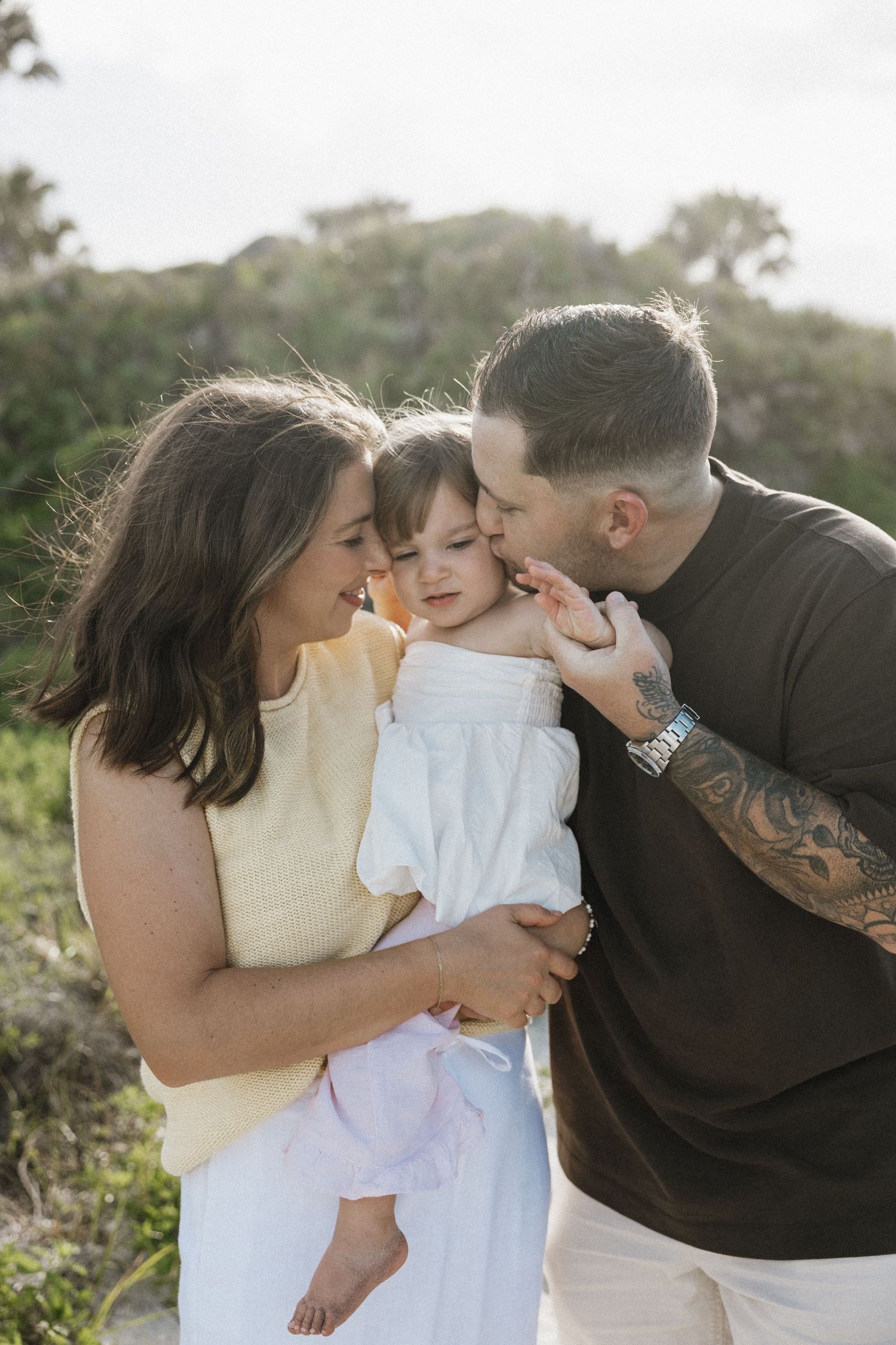Parents lovingly kissing their baby girl outdoors with lush green hillside in background.