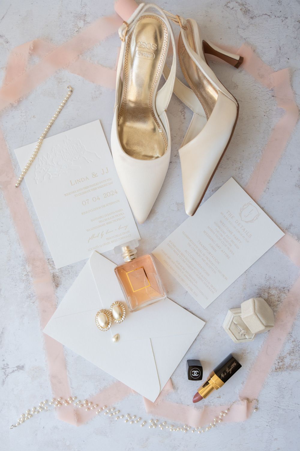 Wedding accessories including white heels, invitations, perfume and jewelry arranged on pink background.