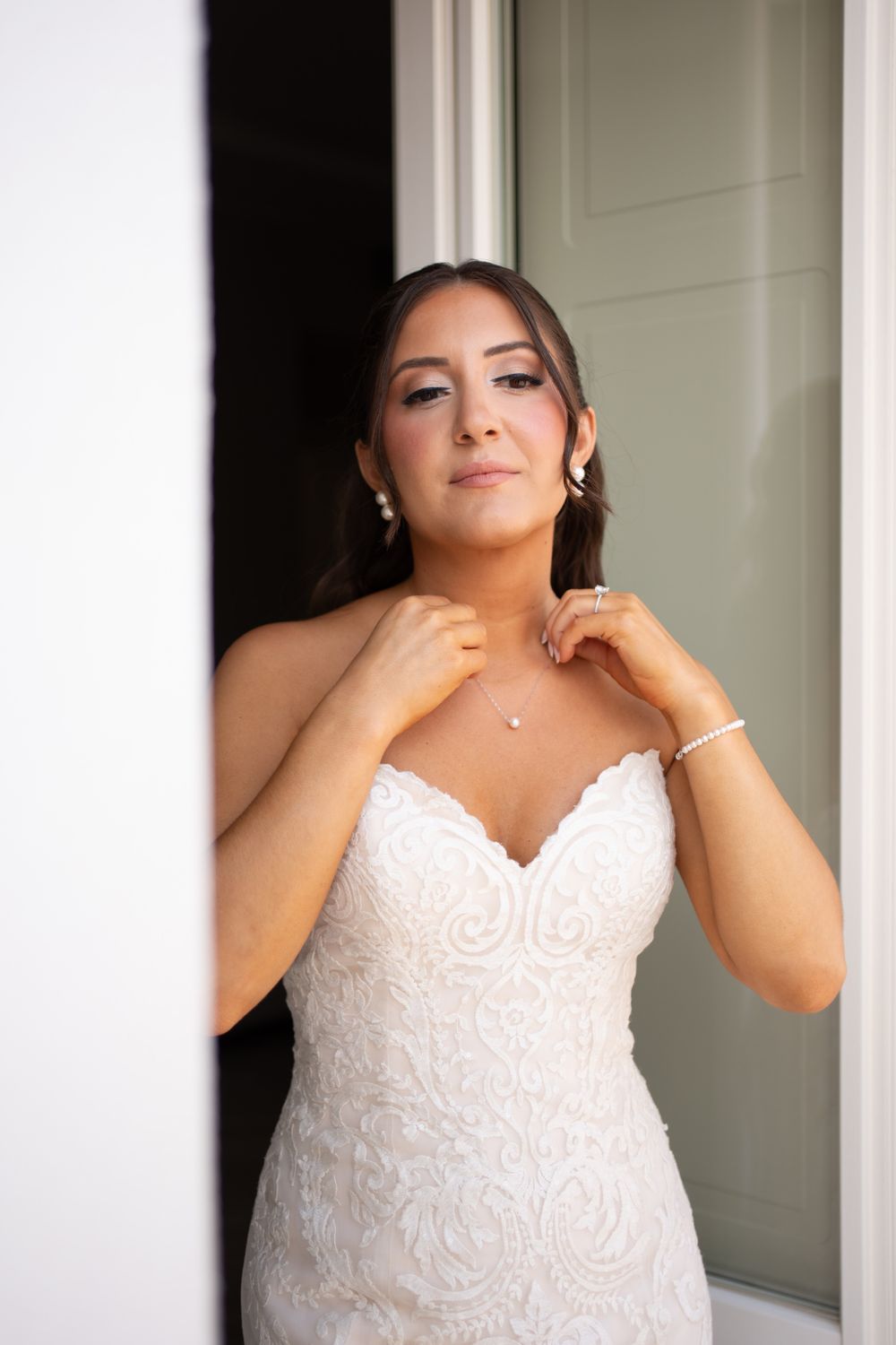Bride in lace wedding gown adjusts jewelry near white doorway.